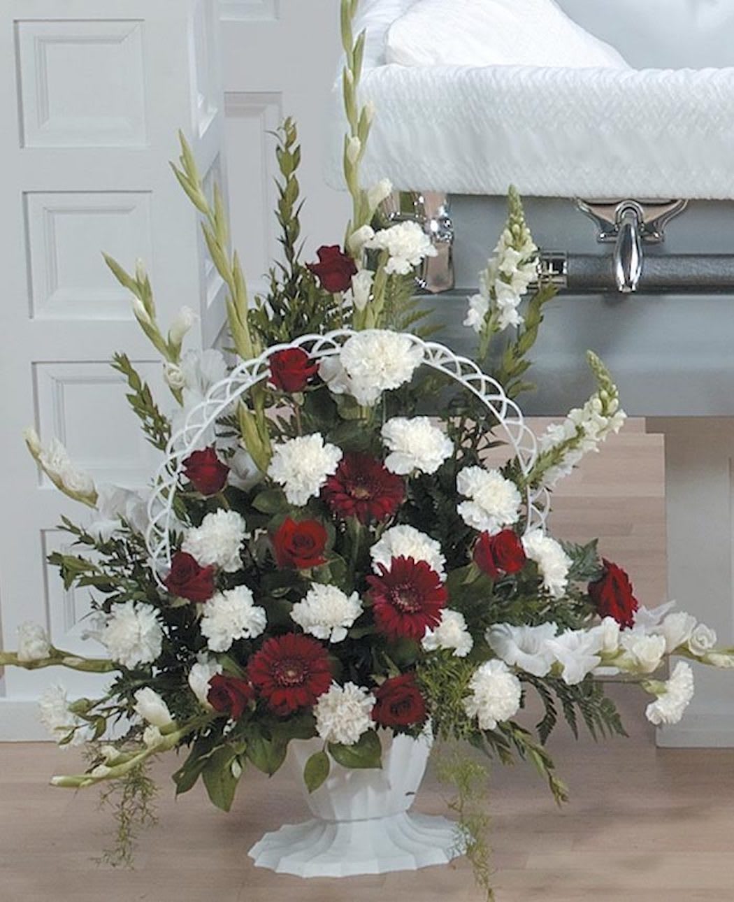 Tall red and white funeral flower arrangement in a white urn beside a casket