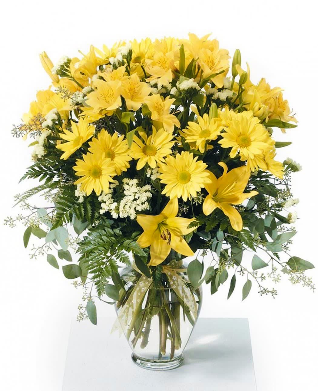 Tall arrangement of yellow lilies and daisies with white accents in a clear glass vase with ribbon