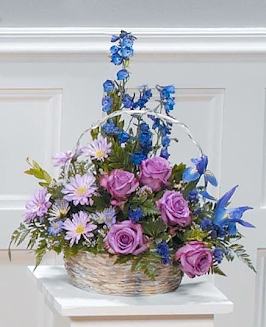 Basket arrangement of purple roses, lavender daisies, and blue flowers with tall blue accents