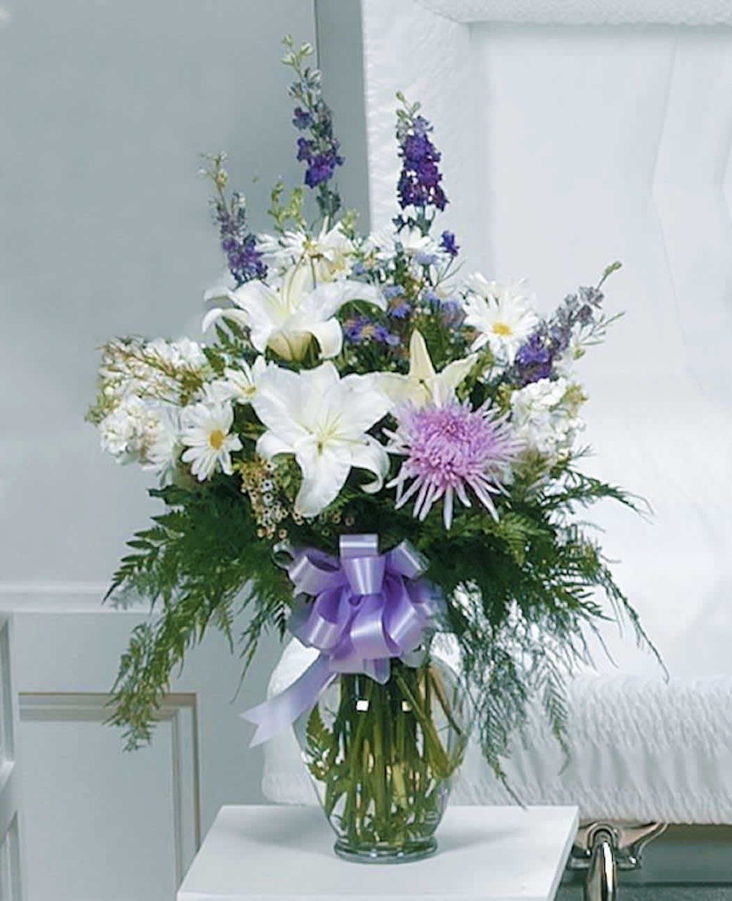 Tall vase arrangement with white lilies, daisies, lavender mums, and purple blooms with a lavender bow.