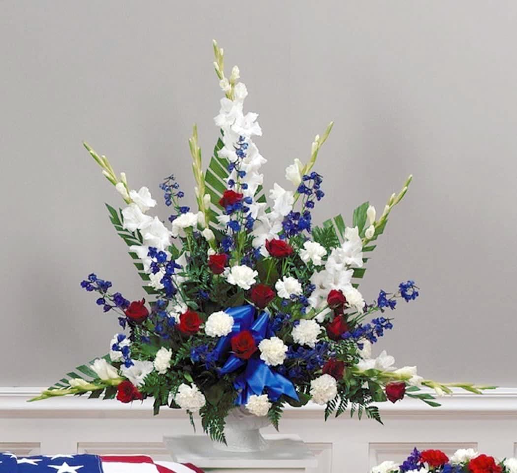 Tall red, white, and blue funeral arrangement with roses, carnations, and blue delphinium in a white pedestal vase