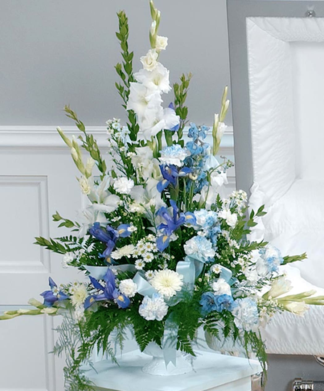 Tall white and blue sympathy flower arrangement in a white pedestal vase beside an open casket