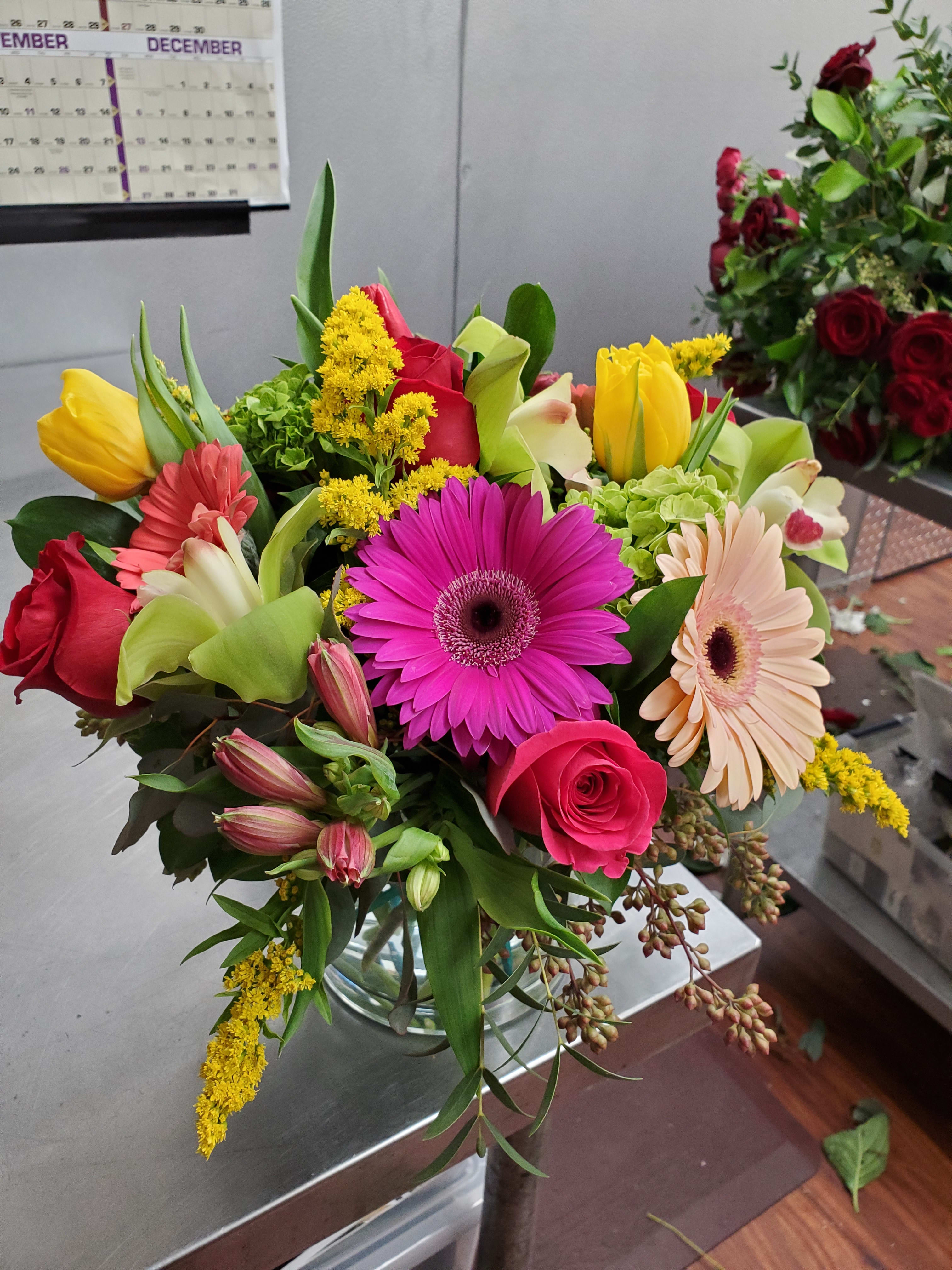 Mixed bouquet of pink gerbera daisies, roses, and yellow tulips in a clear glass vase on a worktable.