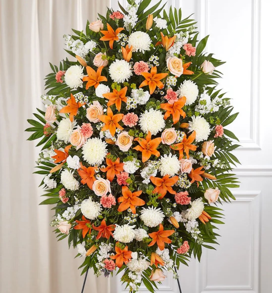 Tall standing spray with orange lilies, white mums, peach roses, and carnations on a leafy easel.