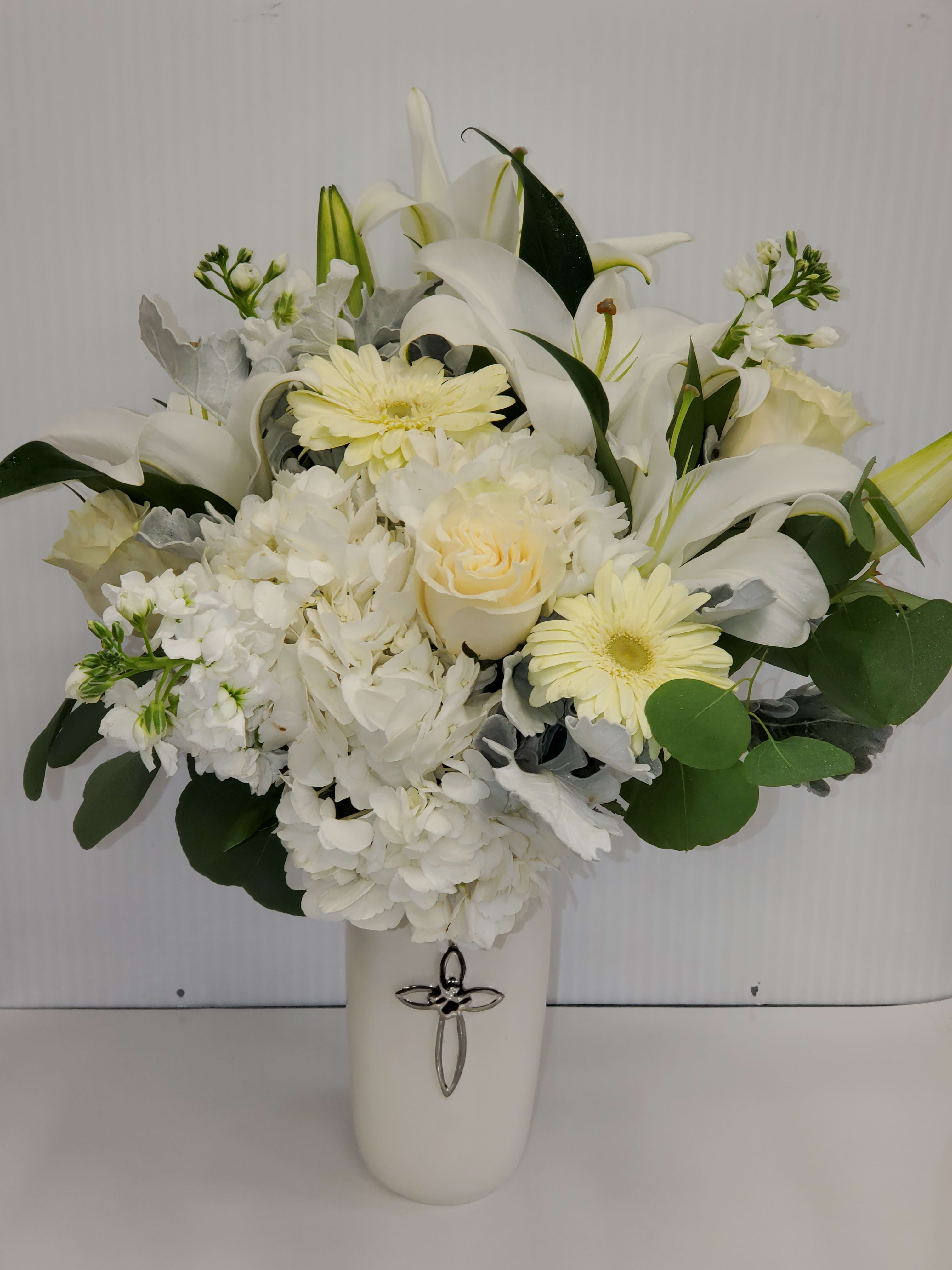 Tall white and cream flower arrangement with lilies, roses, hydrangeas and gerberas in a white vase with a silver cross.