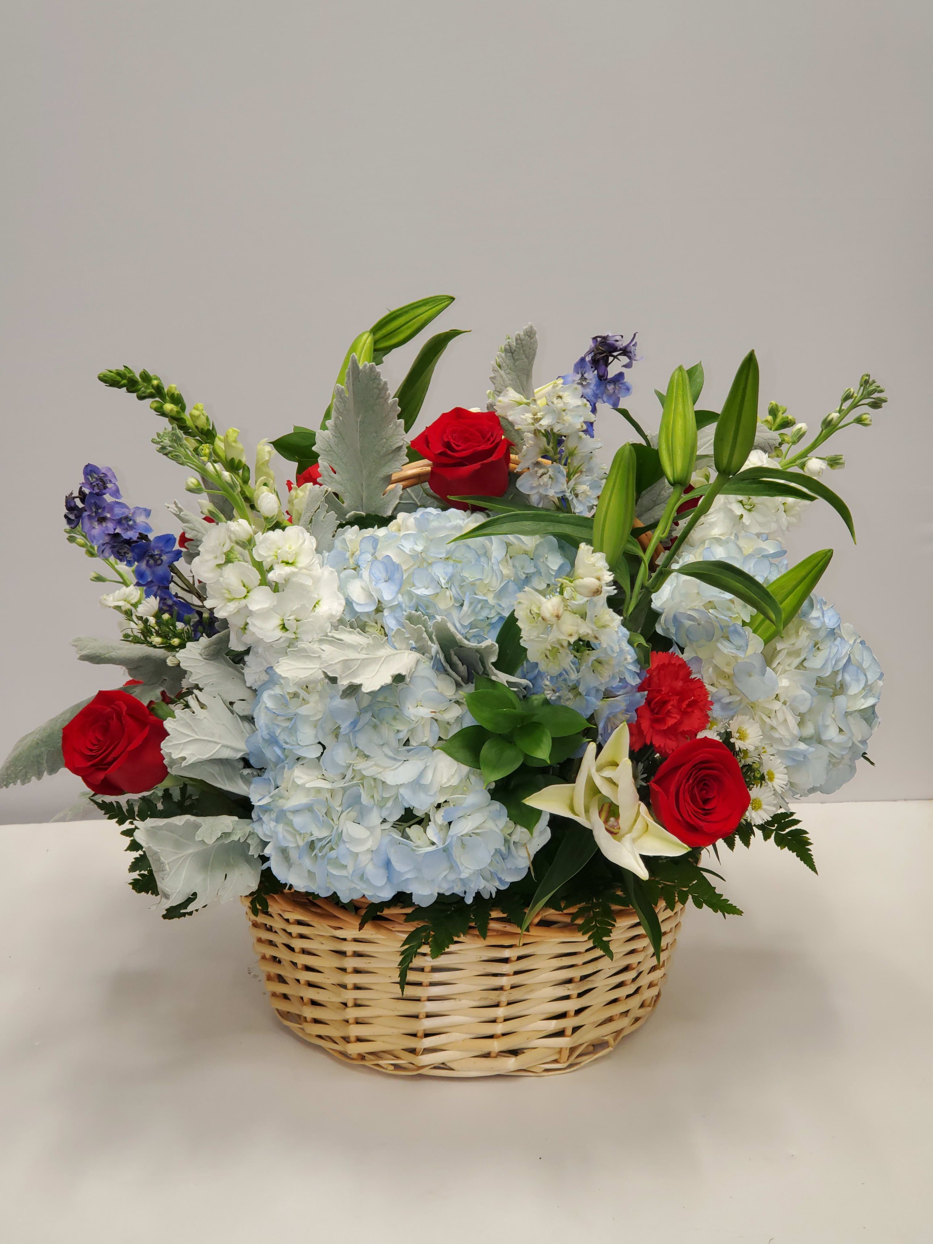 Basket arrangement with blue hydrangeas, red roses, white lilies, and mixed accent flowers