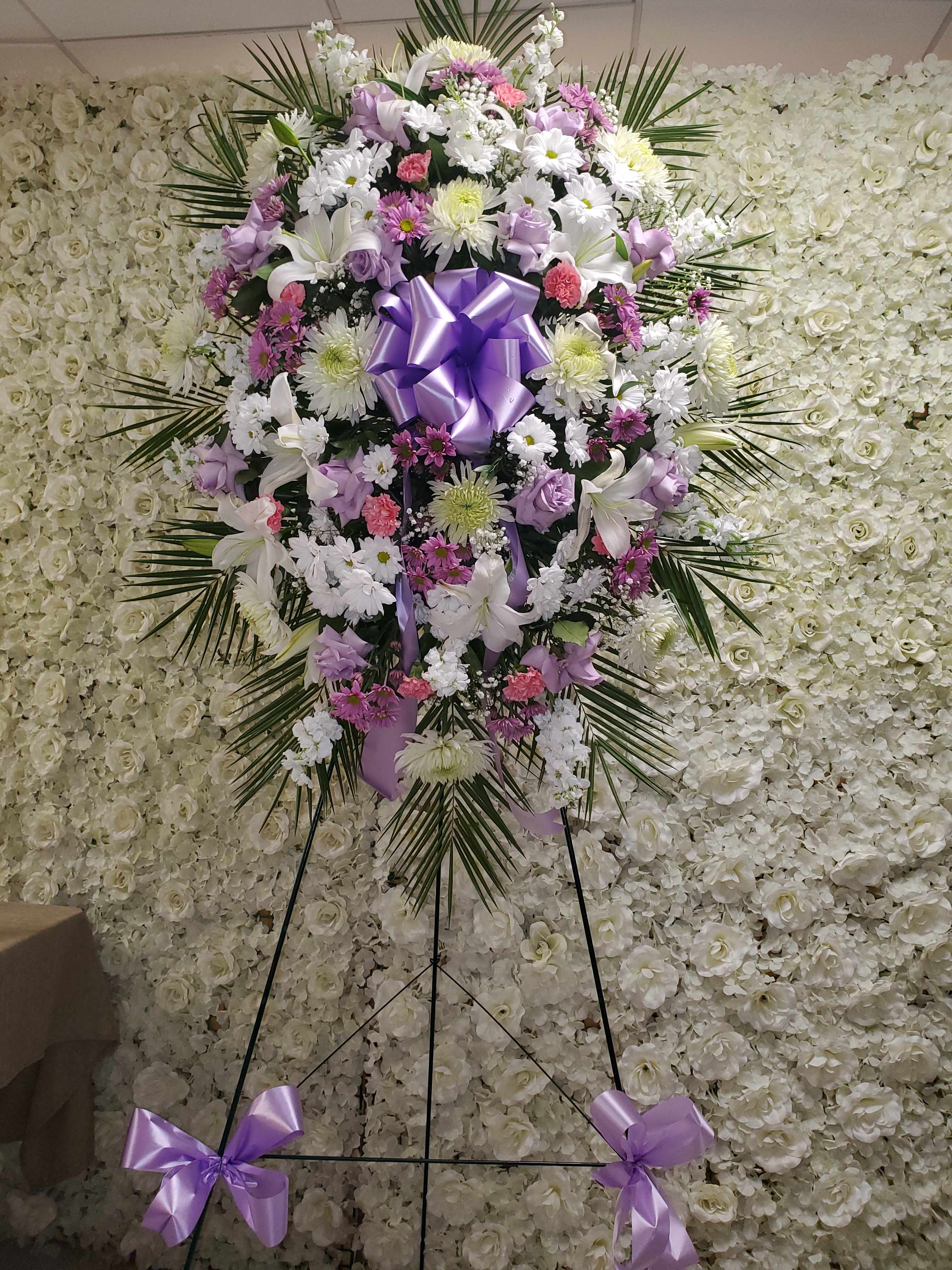 Tall standing spray of white, lavender, and pink flowers with a large lavender bow on an easel.