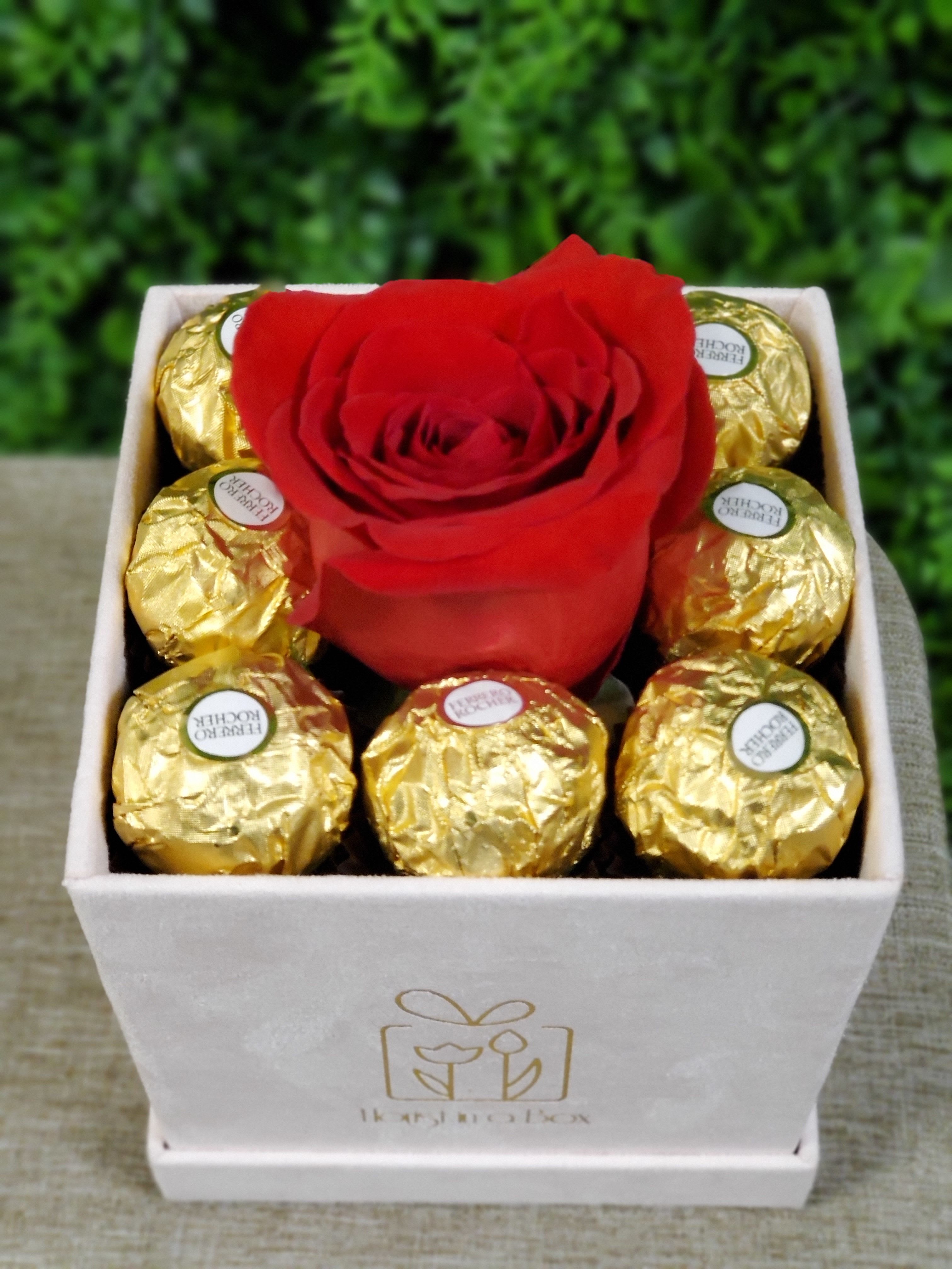 Square box with a single red rose surrounded by gold-wrapped chocolates