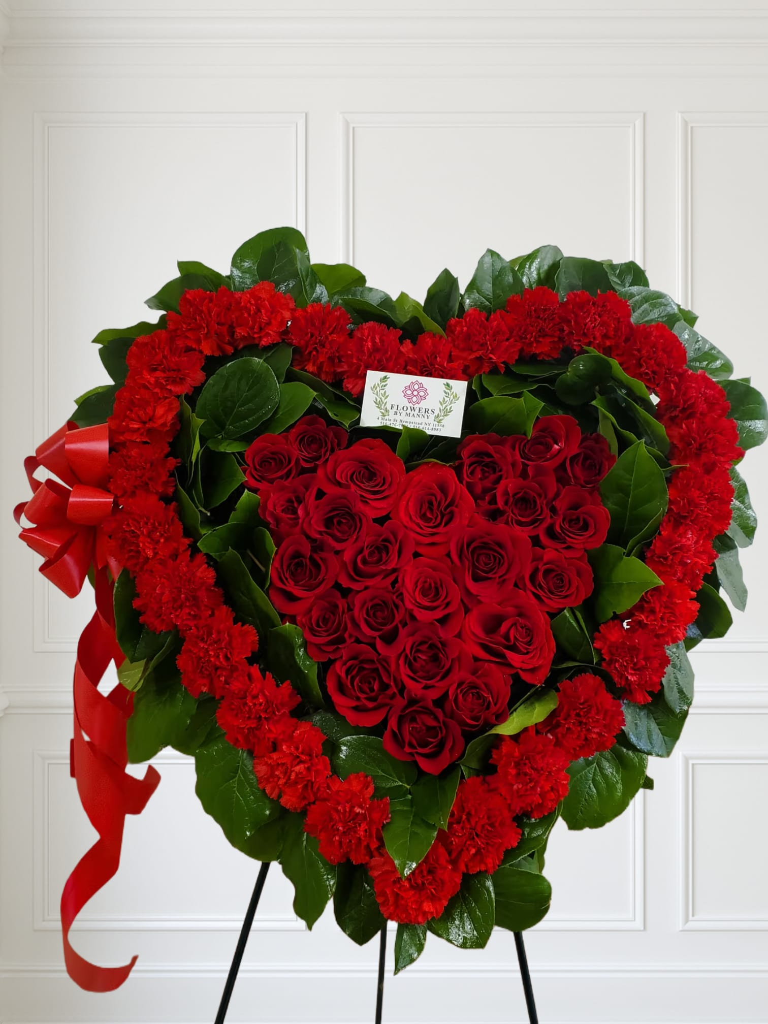 Heart-shaped standing wreath of red roses and carnations with greenery and a red ribbon