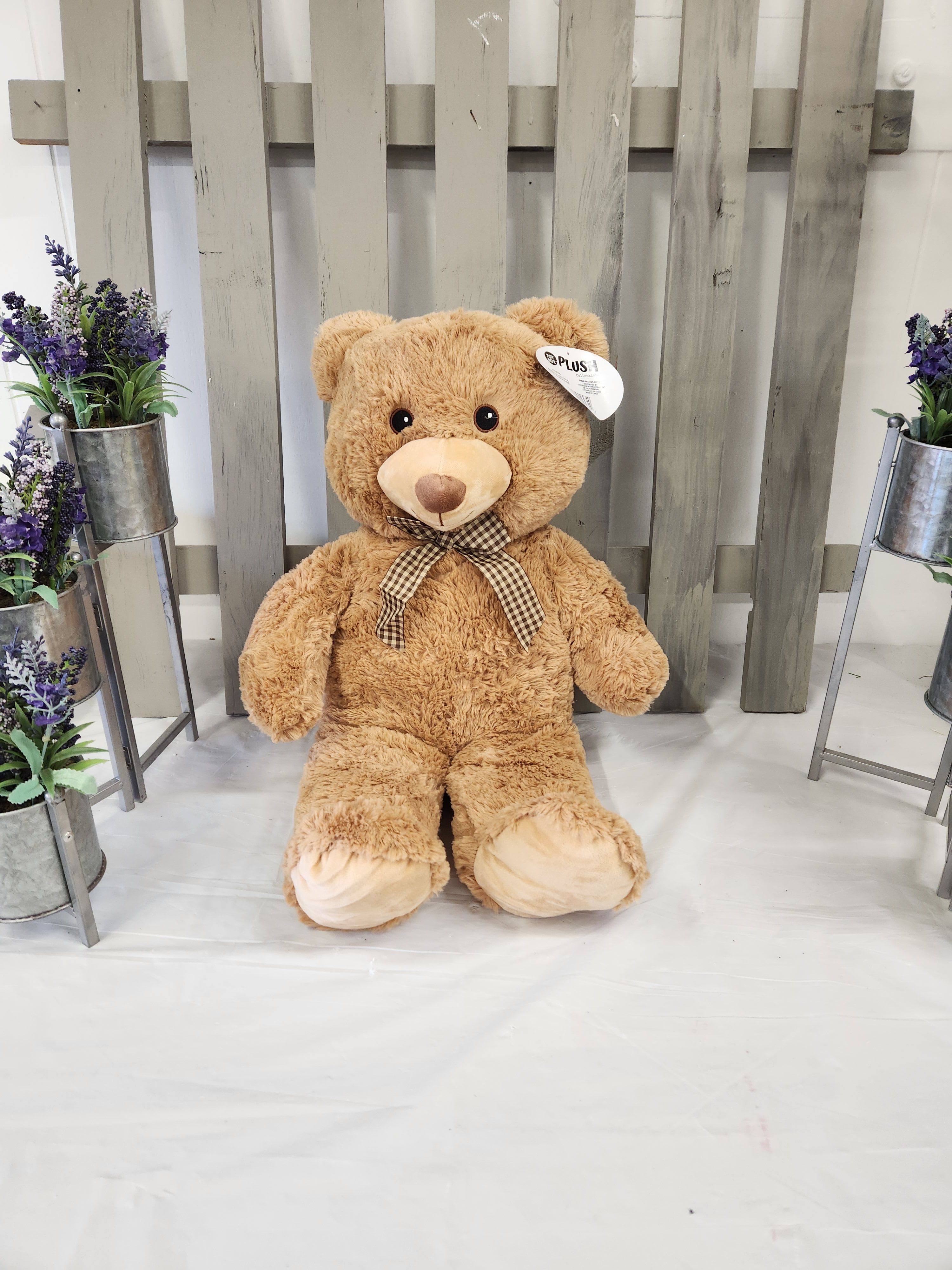 Large teddy bear with a checkered bow, flanked by small purple flower arrangements