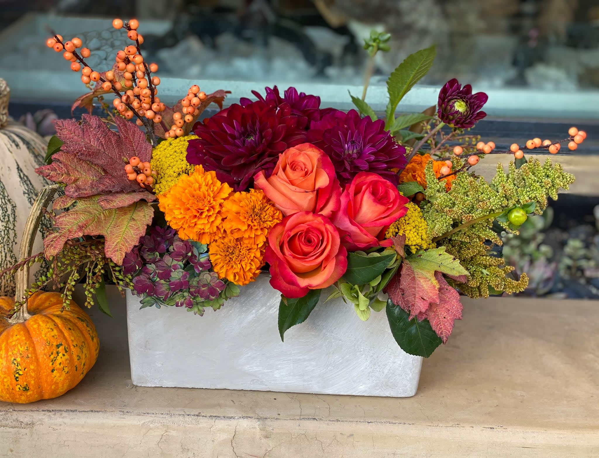 Autumn flower arrangement in a white rectangular container with a pumpkin beside it