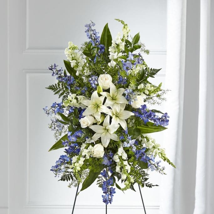 The Faithful Friend Standing Spray - A collection of bold blue flowers shares a tribute that words cannot express. Designed with lilies, roses, delphinium and snapdragons our Faithful Friend™ Standing Spray is a grand gesture to express kindness and solace to a loved one while they are remembering the life they cherished so much.  APPROXIMATE SIZE: 34" H x 24" W. 