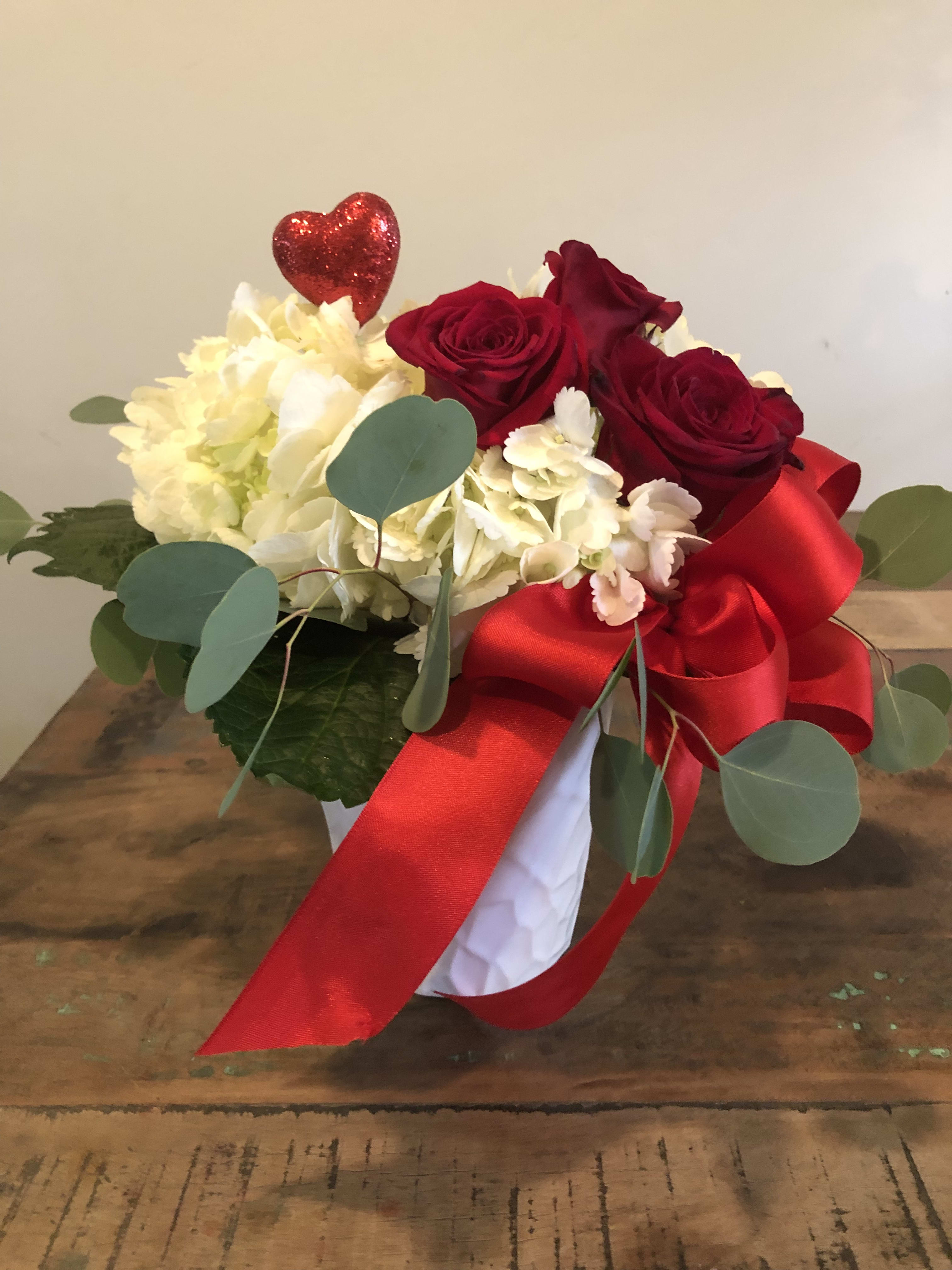 Red roses and white flowers in a white vase with a red ribbon and heart pick