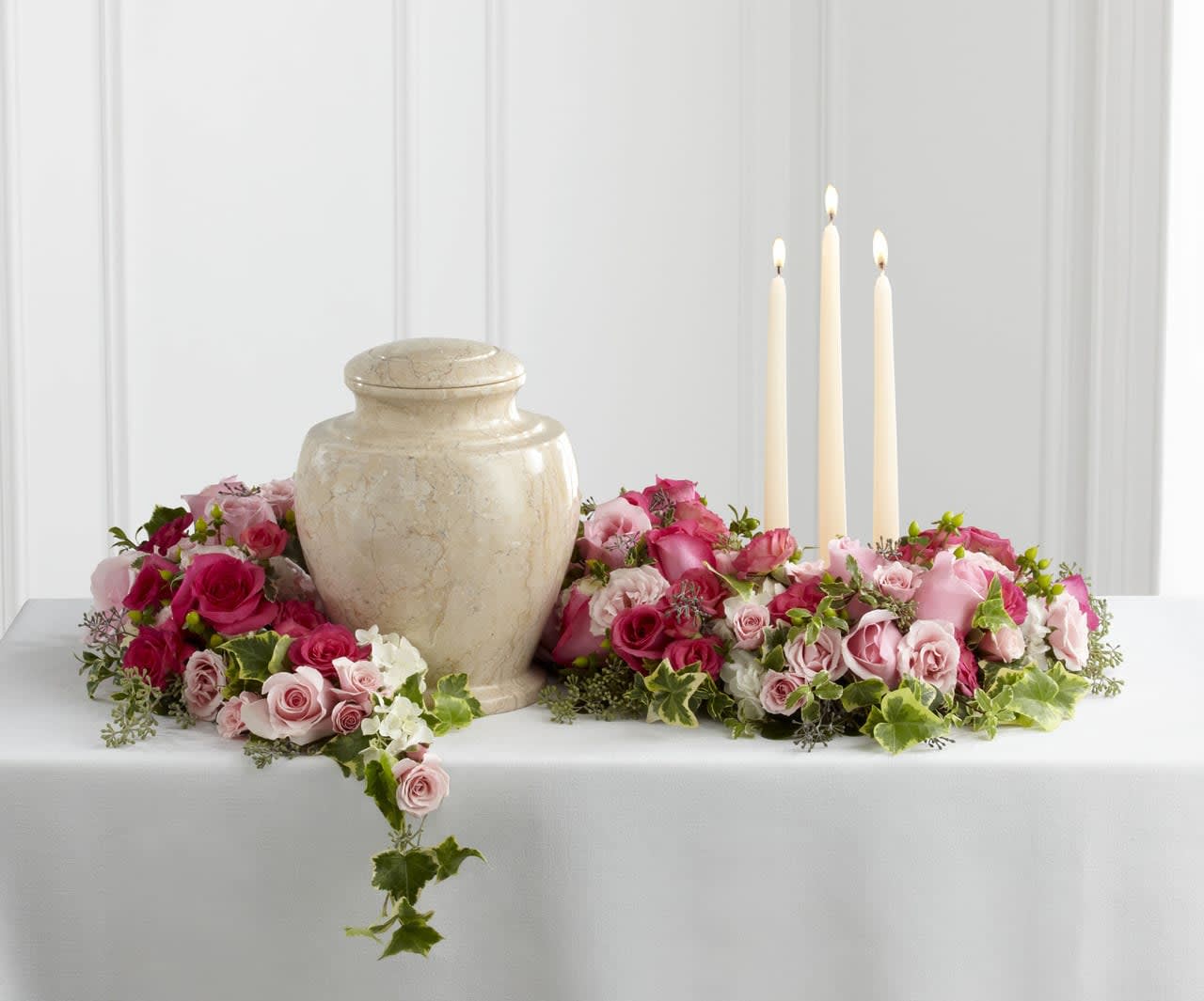 Cremation urn and three ivory candles encircled by pink and white rose flowers on a white table