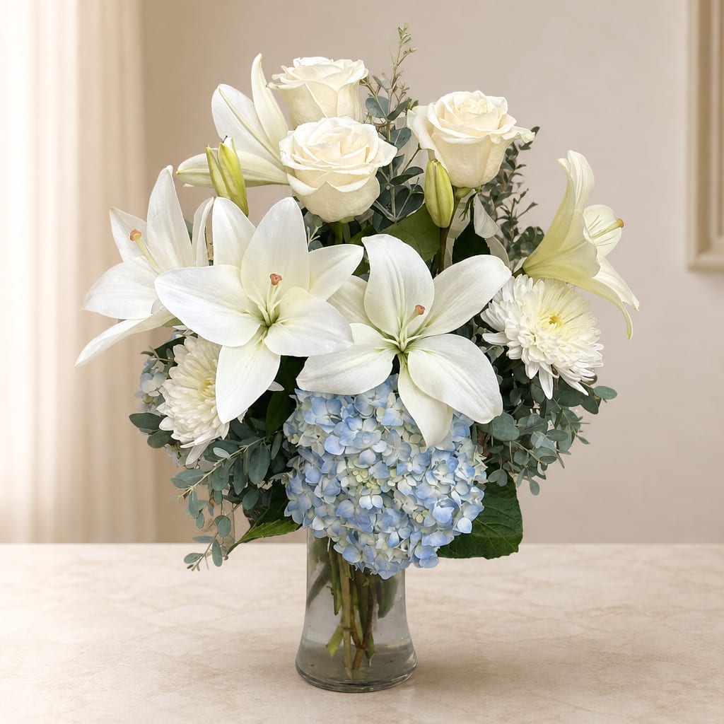 White lilies, roses, and blue hydrangeas arranged in a clear glass vase