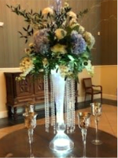 Show Stopper - A luminous centerpiece that commands attention, perfect as wedding flowers for a head table or reception entrance. The Show Stopper pairs soft blue hydrangeas and creamy white roses with additional white blooms and arching greenery to create a ballroom‑worthy display that feels both romantic and grand. Strands of crystal bead garlands catch the light and lend formal sparkle, so the person you're celebrating will feel truly honored the moment they walk in.  Set in a tall, flared glass vase that glows from within, the arrangement offers height and movement-arching foliage and cascading beads draw the eye upward while LED lights inside the vase give the whole piece a warm, steady glow. This design reads beautifully in white, blue, and green tones and is intended to make a memorable statement on round tables, entrance consoles, or behind the head table.  Ideal for weddings where you want an elegant, photo‑ready focal point, the Show Stopper works for formal receptions and romantic ceremonies alike; it's the kind of arrangement that makes the honored couple and their guests feel celebrated. 