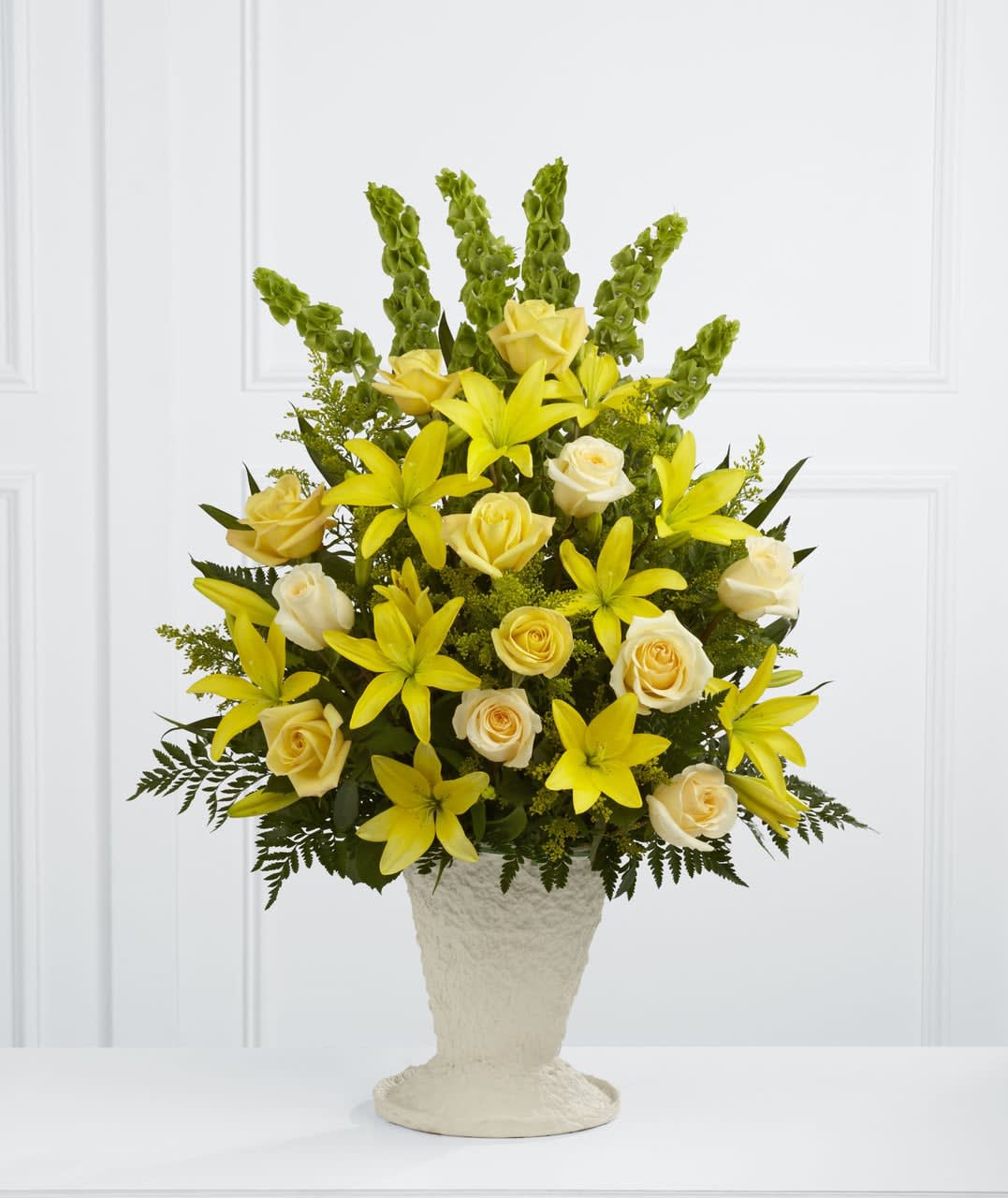 TLSP The Golden Memories Arrangement - The Golden Memories Arrangement bursts with sunlit beauty to honor the life of the deceased. Brilliant yellow Asiatic lilies, roses and solidago are offset by cream roses, Bells of Ireland, teepee palm fronds and lush greens to create a dazzling display. Arranged in a large papier mache urn, this incredible presentation of floral grace will bring a cheerful elegance to their final farewell service. GOOD arrangement includes 19 stems. Approximately 32"H x 21"W. BETTER arrangement includes 24 stems. Approximately 34"H x 23"W. BEST arrangement includes 30 stems. Approximately 36""H x 25"W.  Your purchase includes a complimentary personalized gift message.
