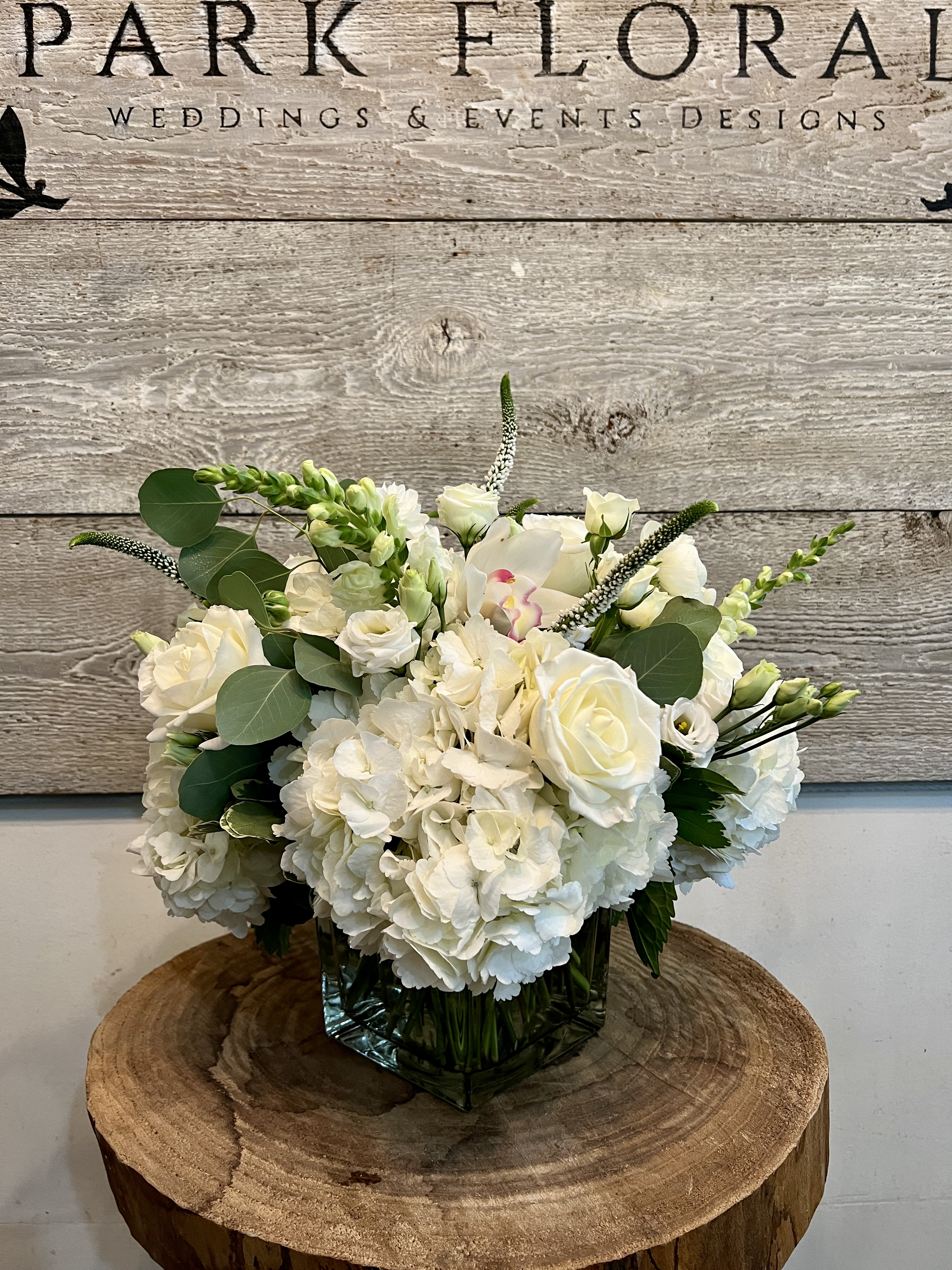 The Park Signature - A lush cluster of white hydrangeas and garden roses creates the first impression: airy, elegant, and perfectly suited as a wedding bouquet, sympathy arrangement, or a refined anniversary gift. This flower arrangement centers on white-and-green tones with soft blush accents, presenting a polished, contemporary look that will make the person you're thinking of feel truly seen and comforted.  The Park Signature combines white roses, full hydrangeas, lisianthus, ranunculus, snapdragons, and a single delicate orchid bloom, all softened by rounded eucalyptus-like foliage and a few upright spikes for graceful height. Arranged in a square glass vase and set on a low wooden pedestal, the composition reads as a medium-sized centerpiece with a dense, rounded form and open, airy texture-classic whites, cool greens, and a whisper of pink for romantic depth.  Perfect for a wedding table, a thoughtful sympathy tribute, or a sophisticated thank-you, this arrangement balances formal presence with gentle warmth. The structured glass vase keeps stems neatly displayed and easy to refresh with water; order now to ensure timely delivery for the occasion you're marking.