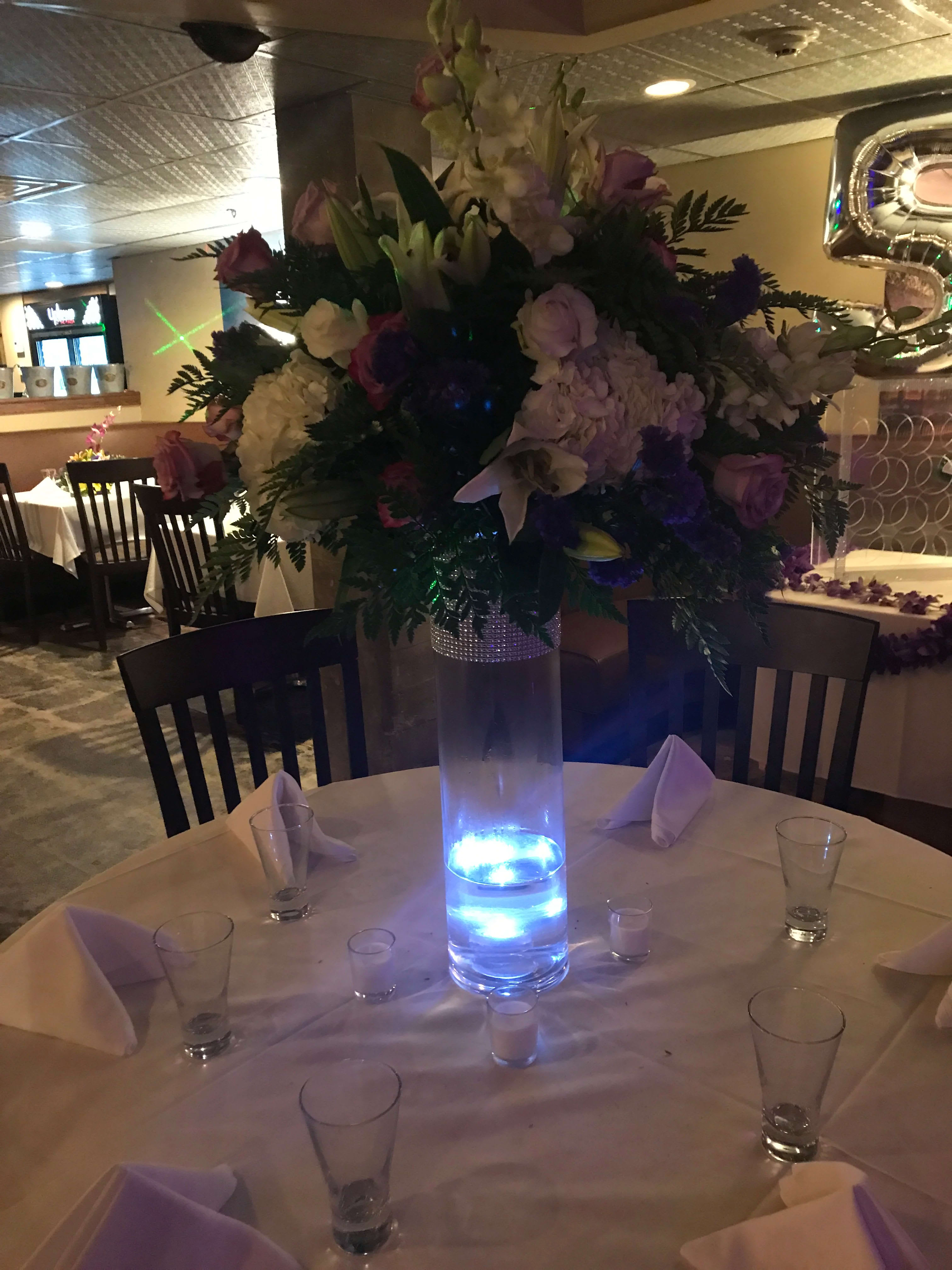 Tall floral centerpiece with pink, white, and purple blooms in a lit glass vase