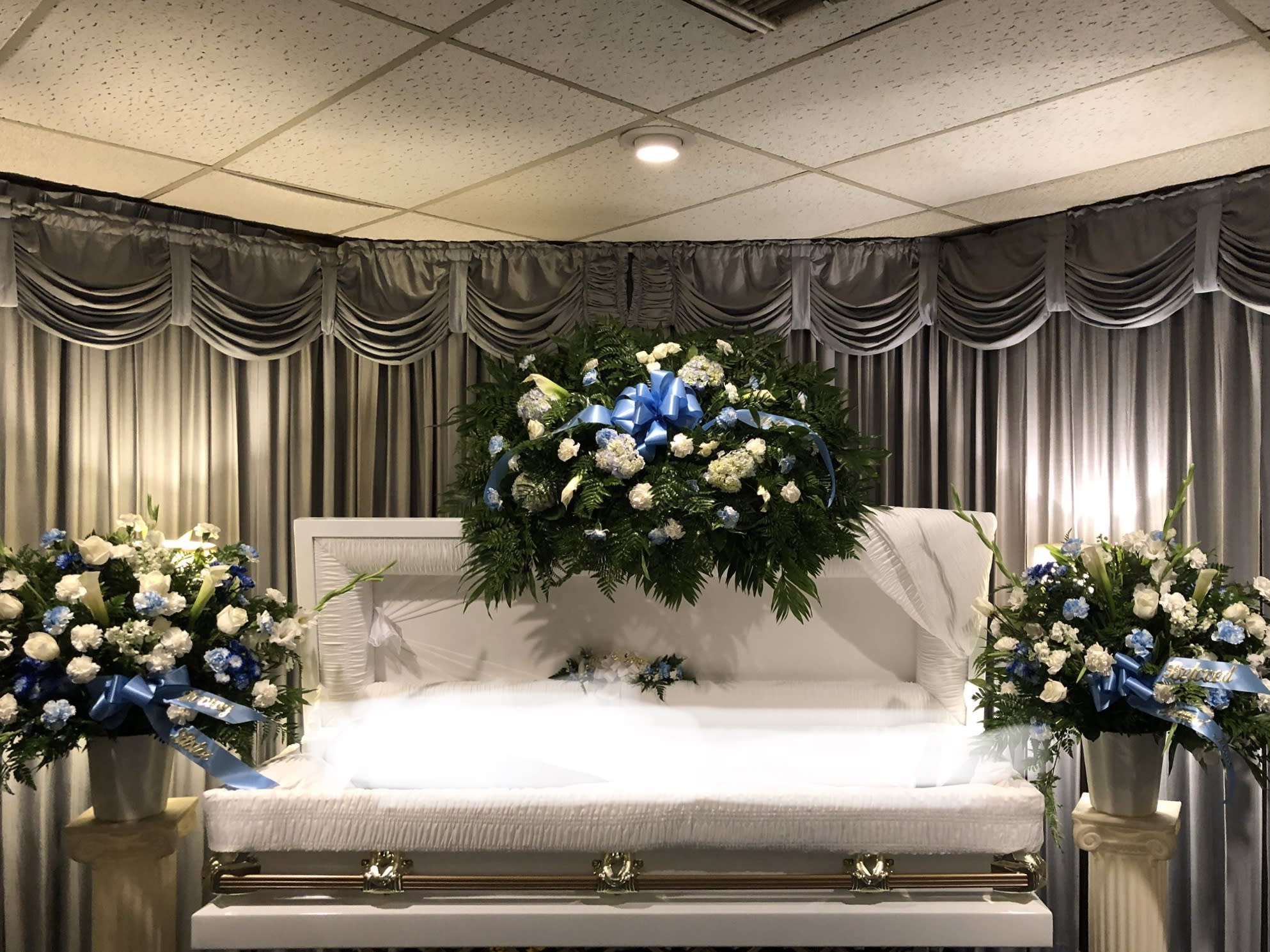 White casket with blue and white floral sprays in a funeral room
