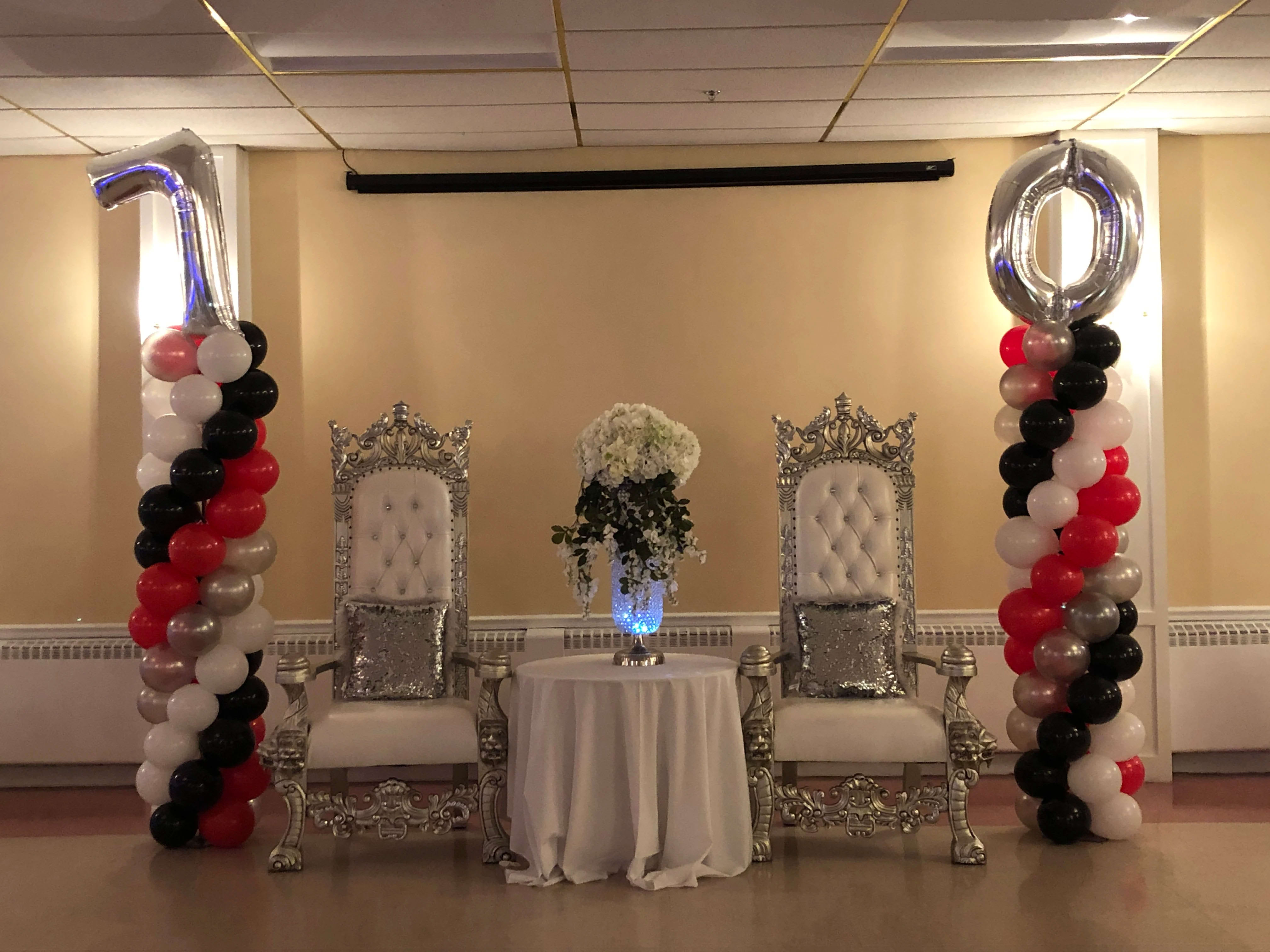 Balloon columns flank two ornate chairs with a floral centerpiece between them.