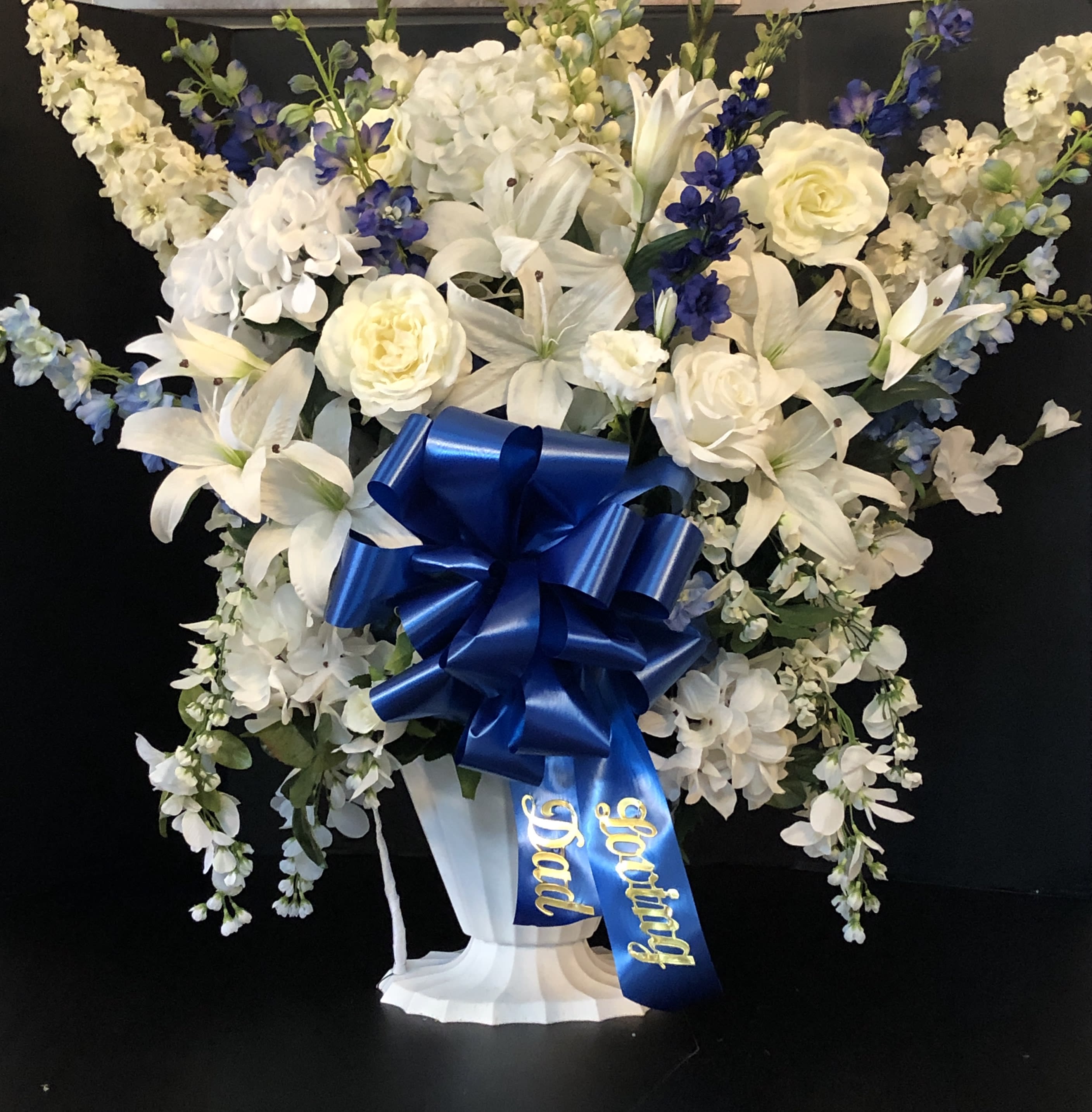 White and blue floral standing arrangement with a large ribbon on a pedestal vase