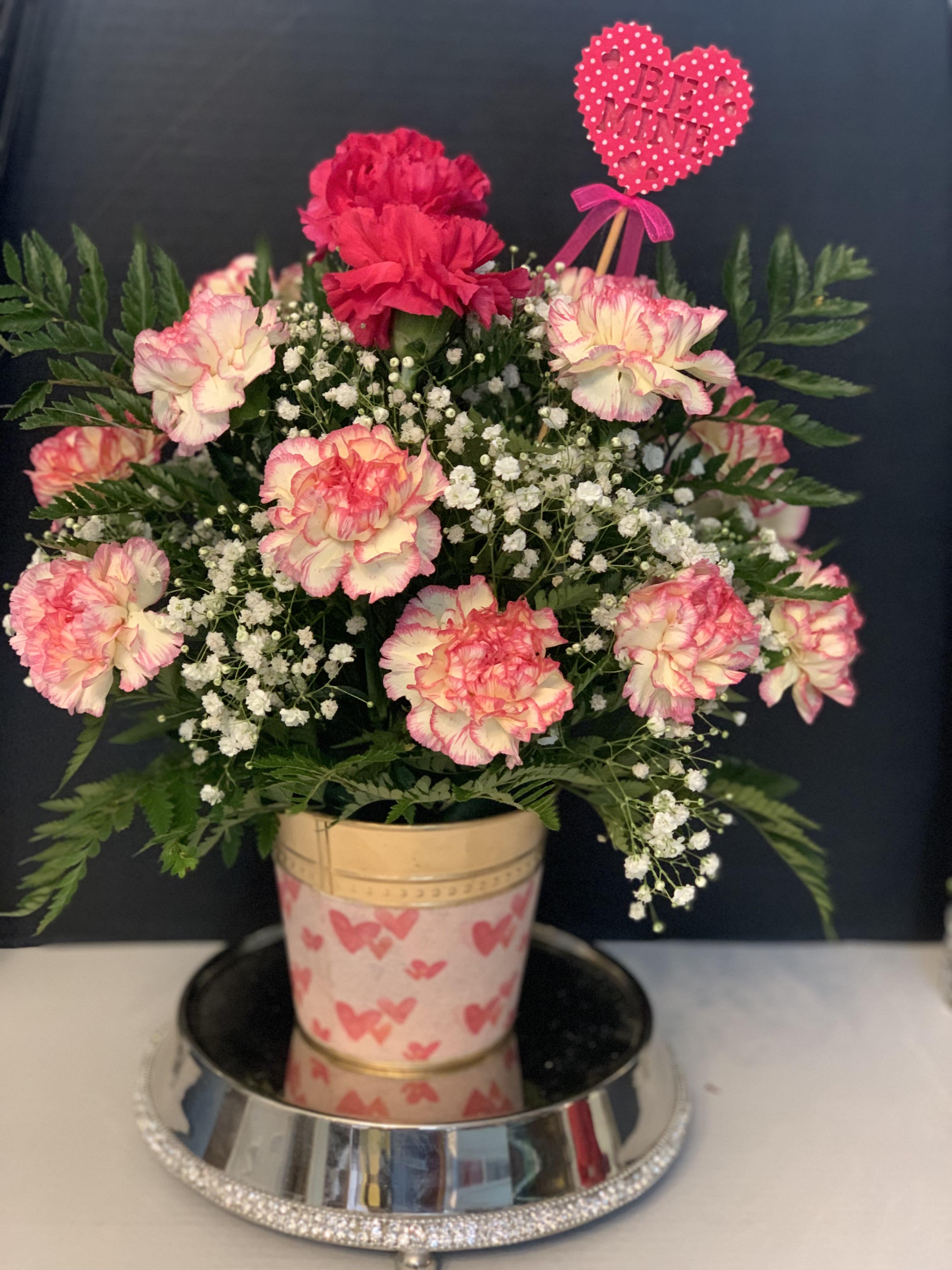 Pink and white carnations in a decorated pot with a heart pick
