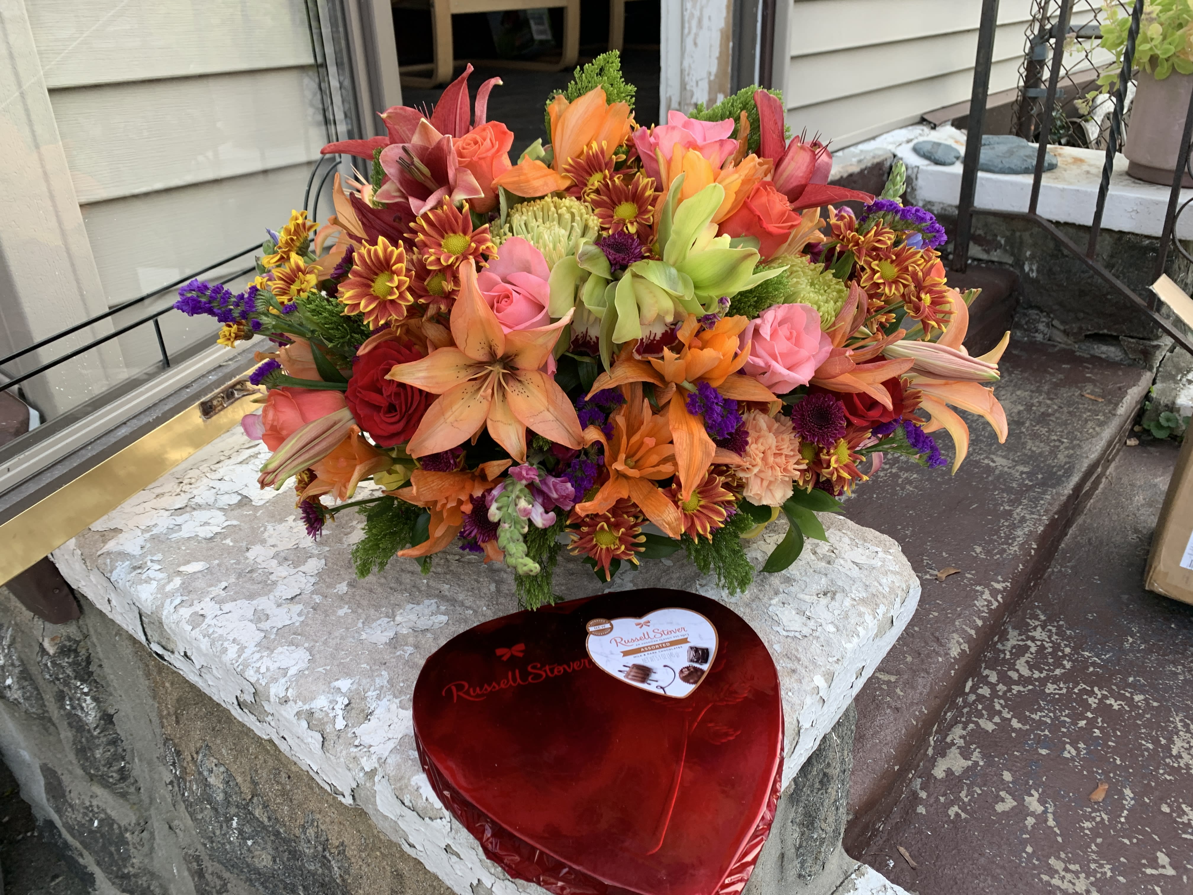 Bright mixed bouquet with roses, lilies, and chrysanthemums beside a heart-shaped chocolate box.