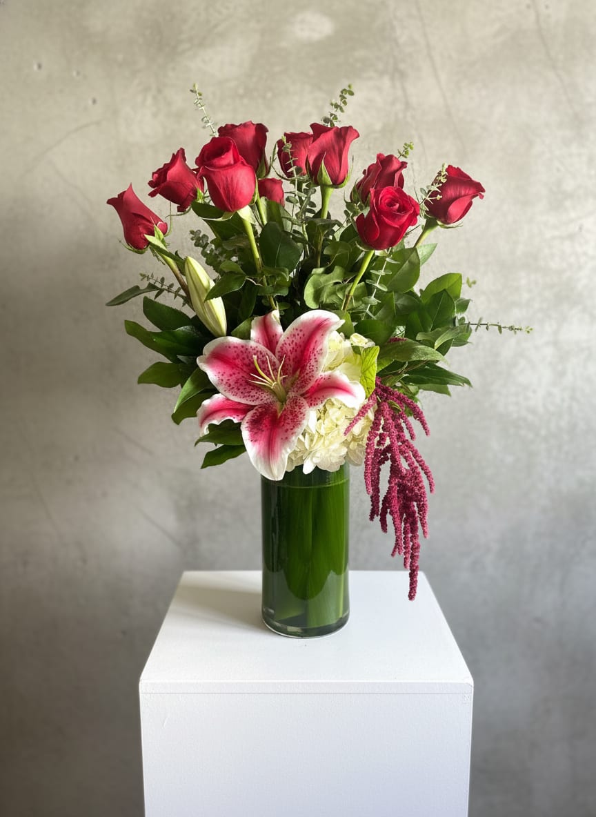Sarah's Roses - Deep red roses pair with a large pink stargazer lily and a cloud of white hydrangea for a romantic, celebratory arrangement that reads like an elegant anniversary or birthday bouquet. Arranged upright in a tall green glass vase, the mix of red, pink, white and green - with trailing burgundy/magenta amaranthus and broad foliage - creates a full, layered silhouette that feels both formal and heartfelt. The color contrast is striking enough for Valentine's Day or prom, yet softened by white blooms so it also works for "just because," congratulations, or a thoughtful get-well gift.  This arrangement includes a dozen standing red roses, a prominent pink-and-white lily, a pale hydrangea cluster, and hanging amaranthus set in a cylindrical green glass vase; the stems sit upright to give height and presence on a table or mantle. The texture of the hydrangea balances the sleek roses and the lily's fragrance, while the trailing blooms add graceful movement. Ideal for anniversaries, graduations, Mother's Day, or to say "I'm sorry" or "thank you," it arrives ready to display - minimal care: fresh water, a clean vase, and occasional recut stems will keep the bouquet looking its best.