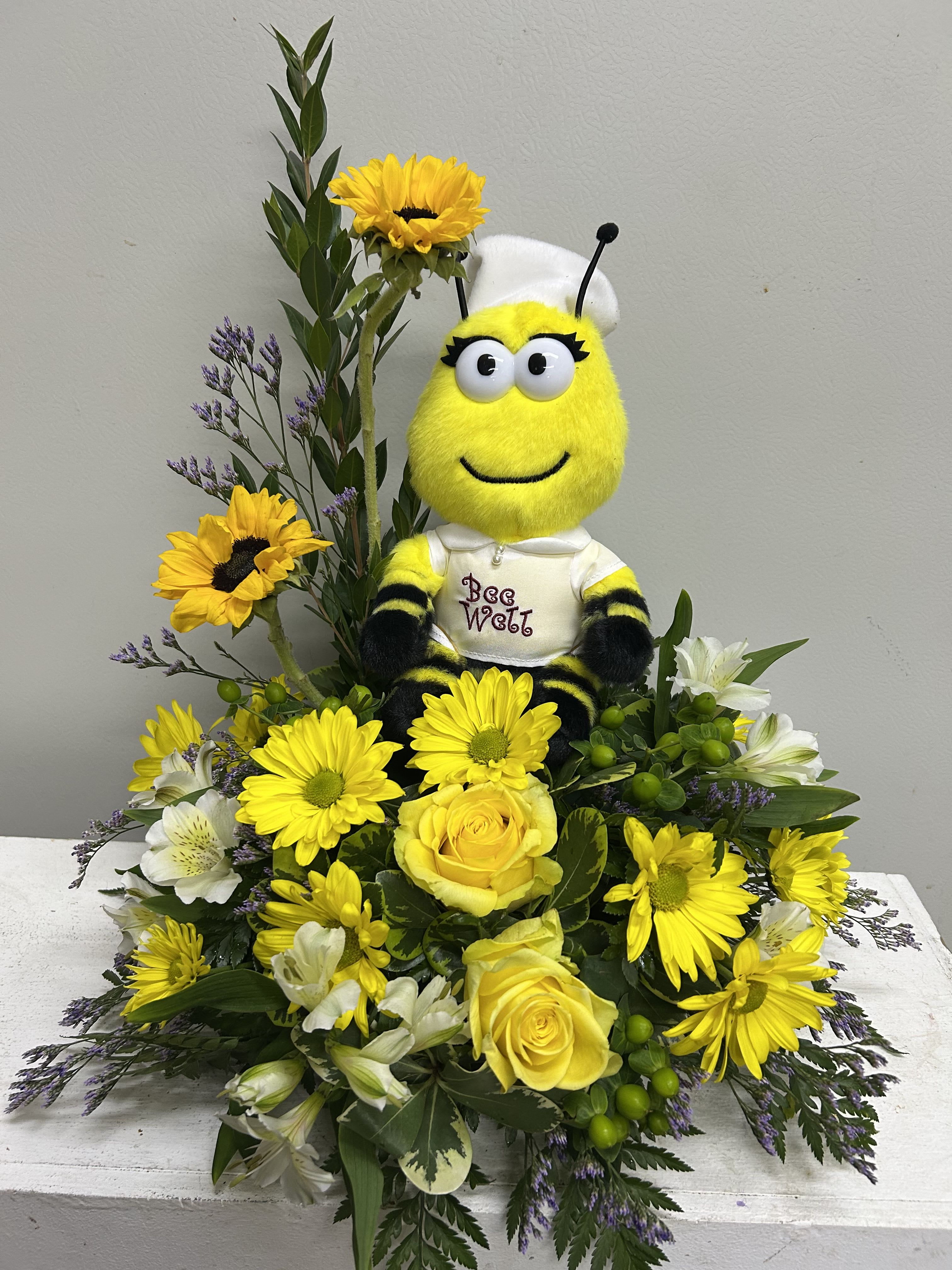 Bee Well - This full, layered design pairs bold yellow sunflowers and roses with white alstroemeria and small purple filler flowers for contrast, all arranged to spread outward around the bee at the center. A soft plush bee wearing a white fabric hat and a shirt that says "Bee Well" sits tucked into a decorative container, giving the piece a playful, comforting personality. The combination of textures-velvety rose petals, crisp alstroemeria, and the soft toy-creates visual depth and a tactile charm recipients will notice.  Send this to a friend or family member recuperating from illness, a new parent needing brightness, or anyone who could use a sunny pick-me-up; she'll feel genuinely seen and encouraged. Vase and sizing are arranged to showcase the plush bee amid the blooms; to keep flowers looking fresh, trim stems and refresh water every 2-3 days.