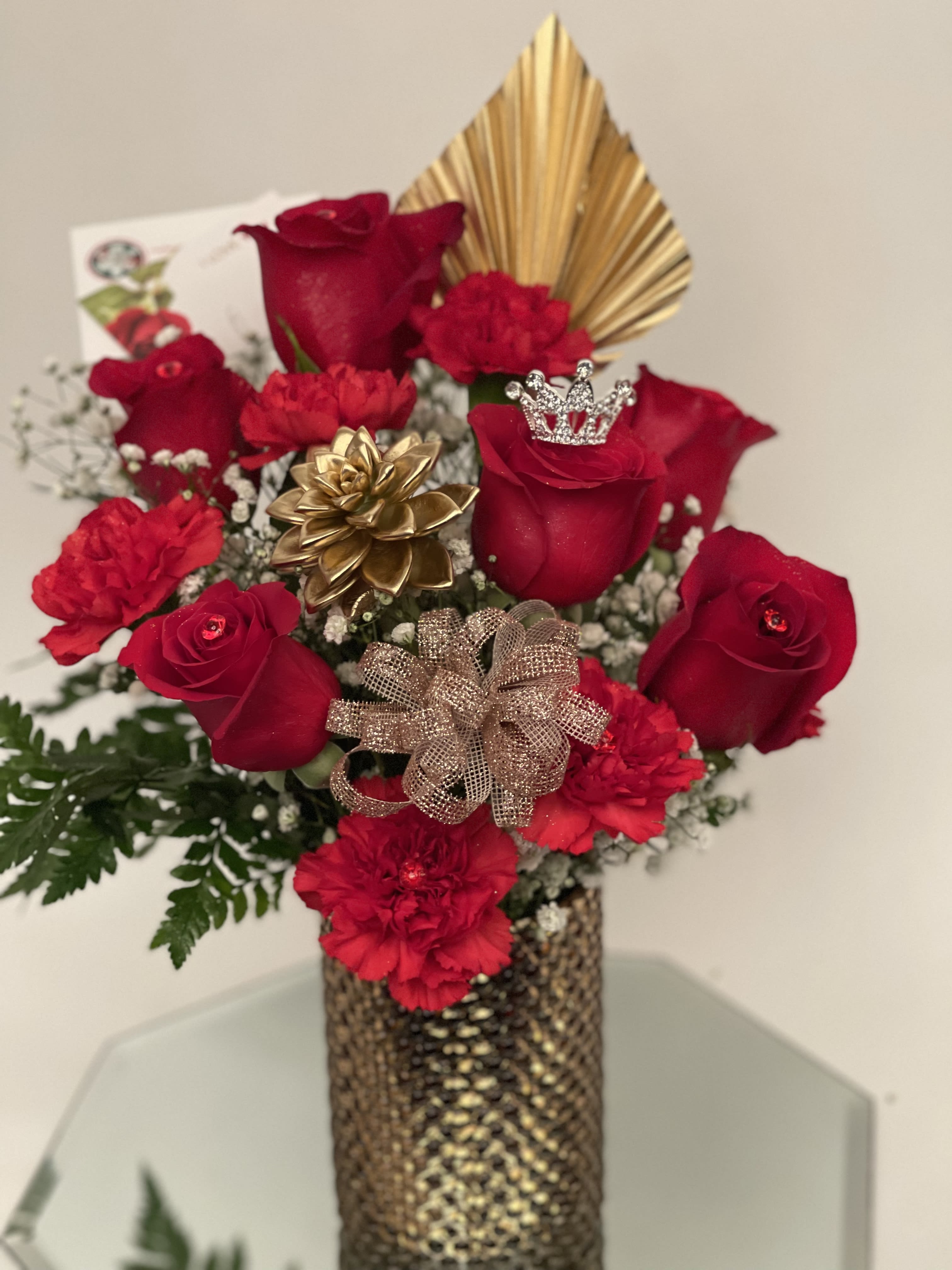 Red roses and carnations in a gold vase with decorative bows and a crown