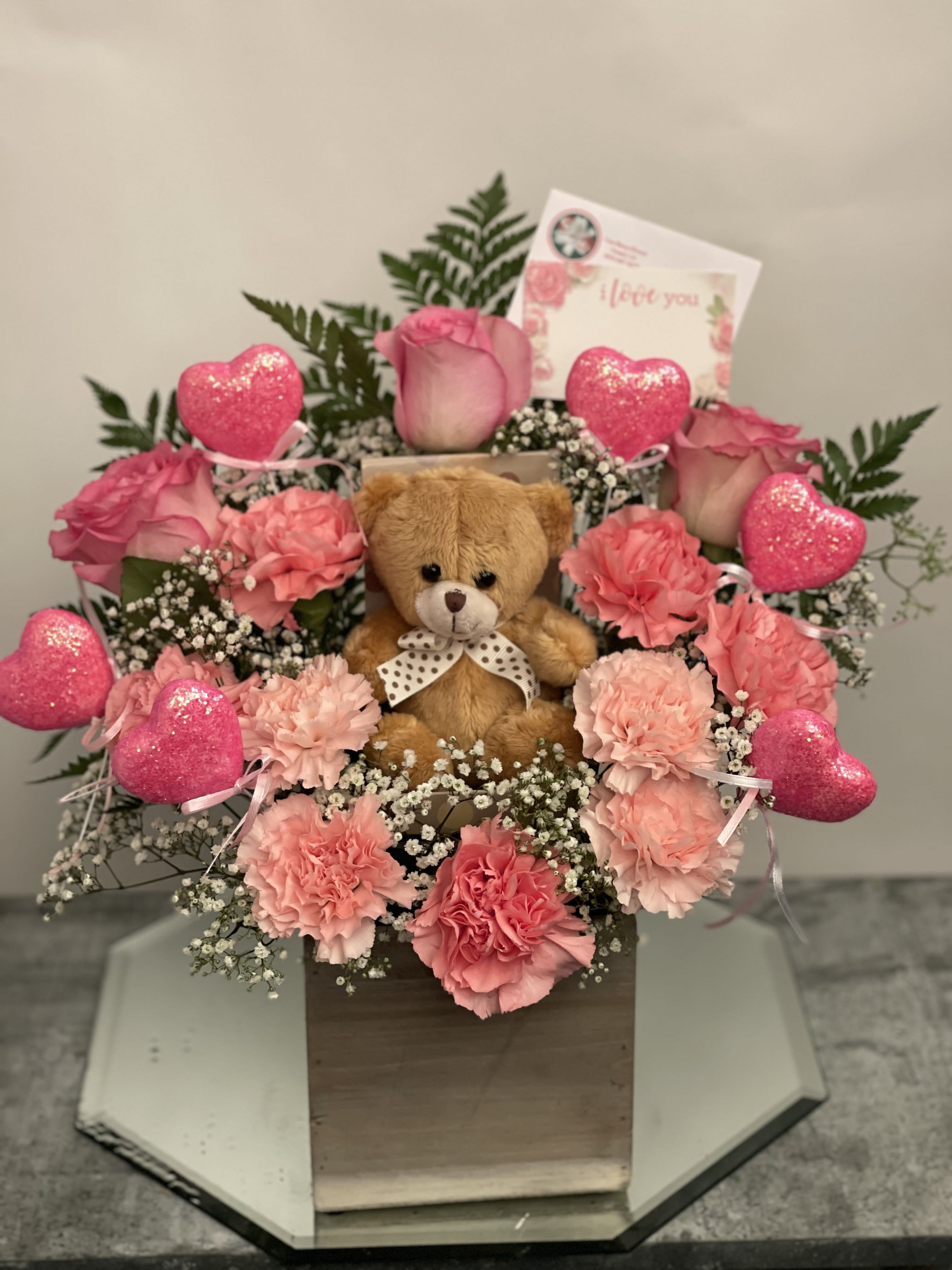 Pink carnations and roses arranged with a teddy bear and heart decorations