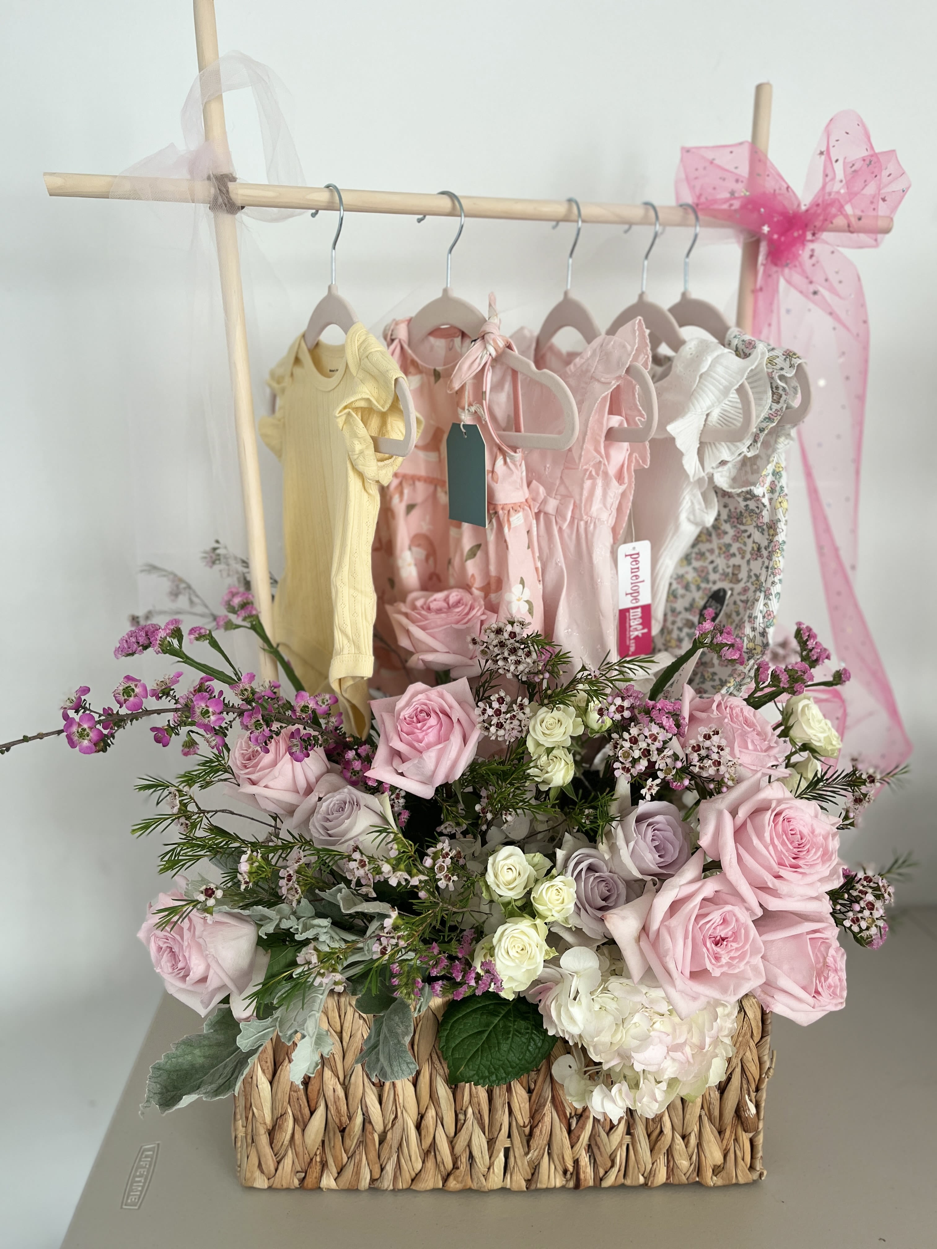 Pink and white rose arrangement in a woven basket with baby dresses behind it