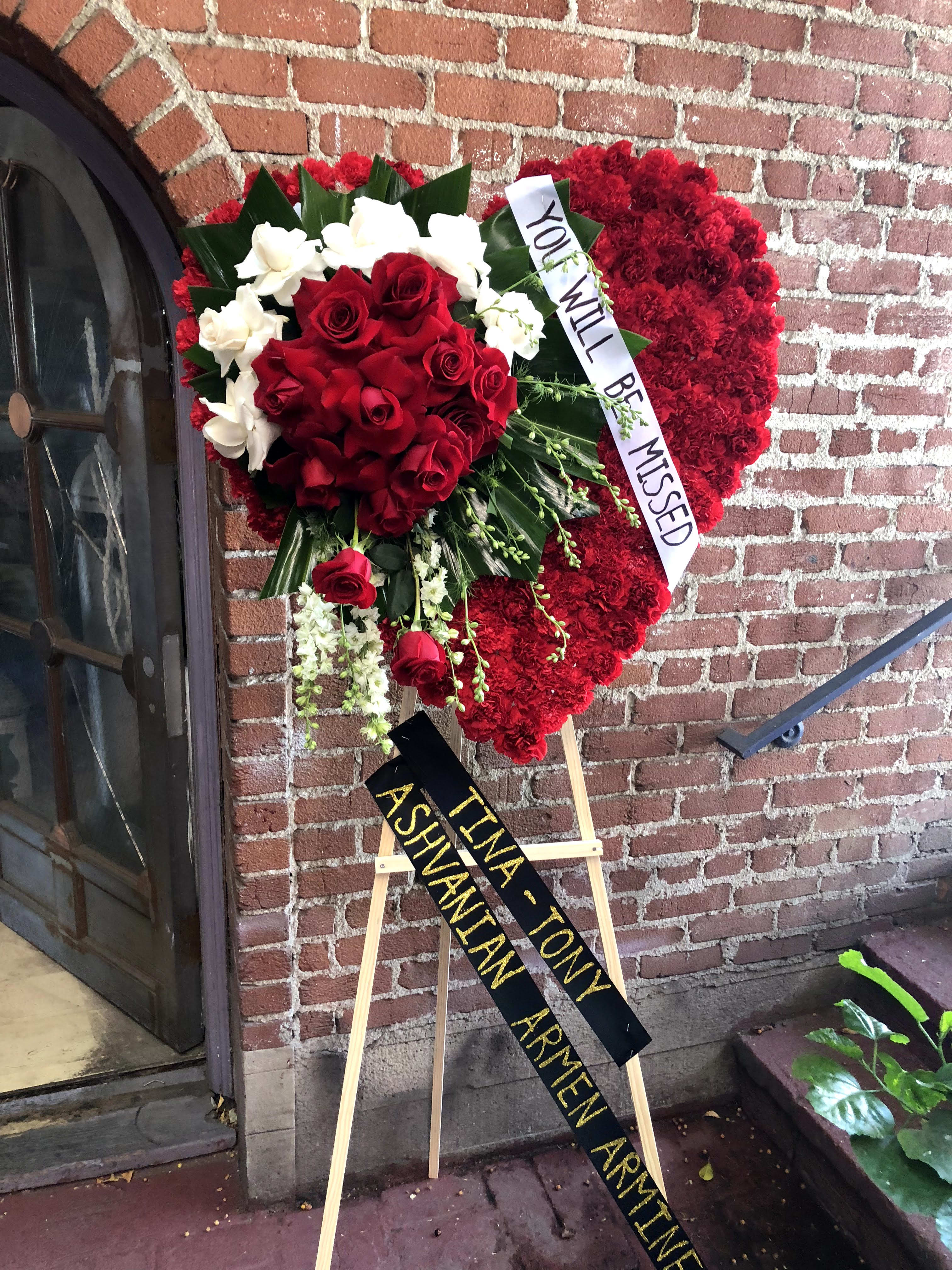 Heart-shaped standing funeral arrangement of red and white roses with memorial ribbons on an easel.
