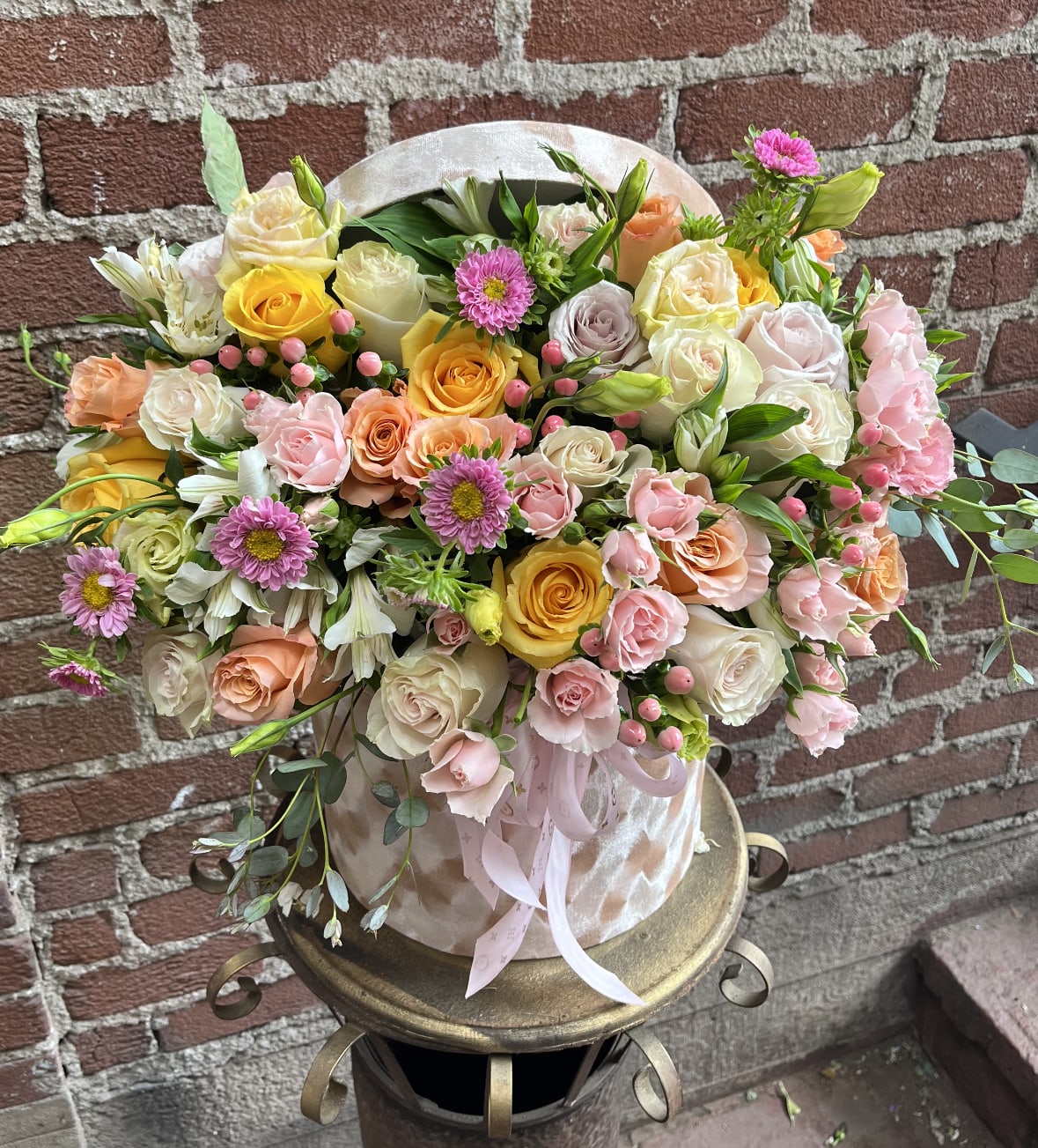 Pastel arrangement of roses, spray roses and daisylike blooms in a pink hatbox-style container