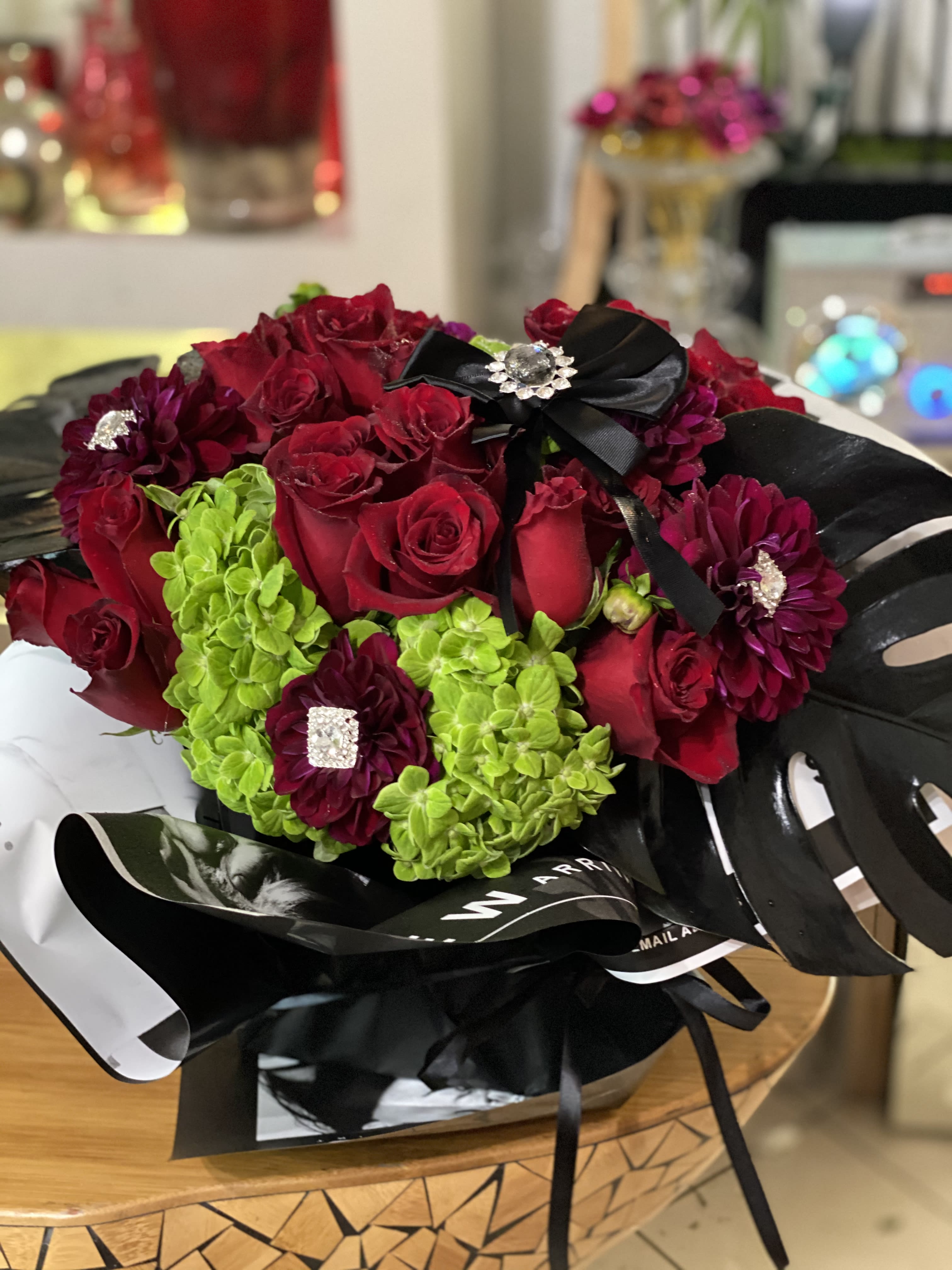 Gothic Red Rose Arrangement  - Wrapped, black, square vase flowing with luscious, red roses. Surrounded with black leaves and ribbons enhanced with shimmering brooches.
