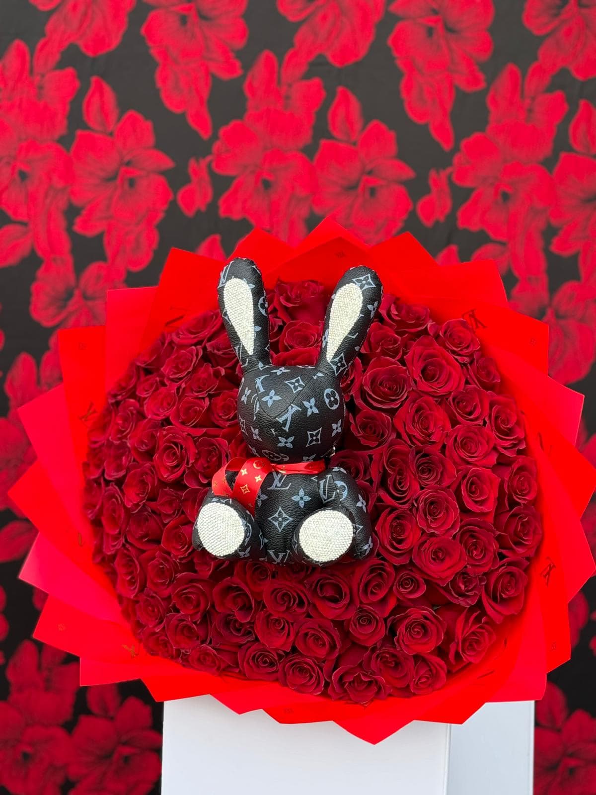 Glamour in Bloom: Red Rose Bouquet with Bunny Accent - This show-stopping bouquet combines the timeless beauty of 100 vibrant red roses with the playful elegance of a Louis Vuitton-inspired bunny with ears and feet all encrusted with dazzling rhinestones. The rich, velvety petals of the roses create a stunning contrast with the high-fashion details, making this piece an unforgettable statement of style and luxury. Perfect for any occasion that calls for a bold, glamorous touch, this bouquet merges romance and opulence, making it an extraordinary gift or centerpiece that will captivate and awe.