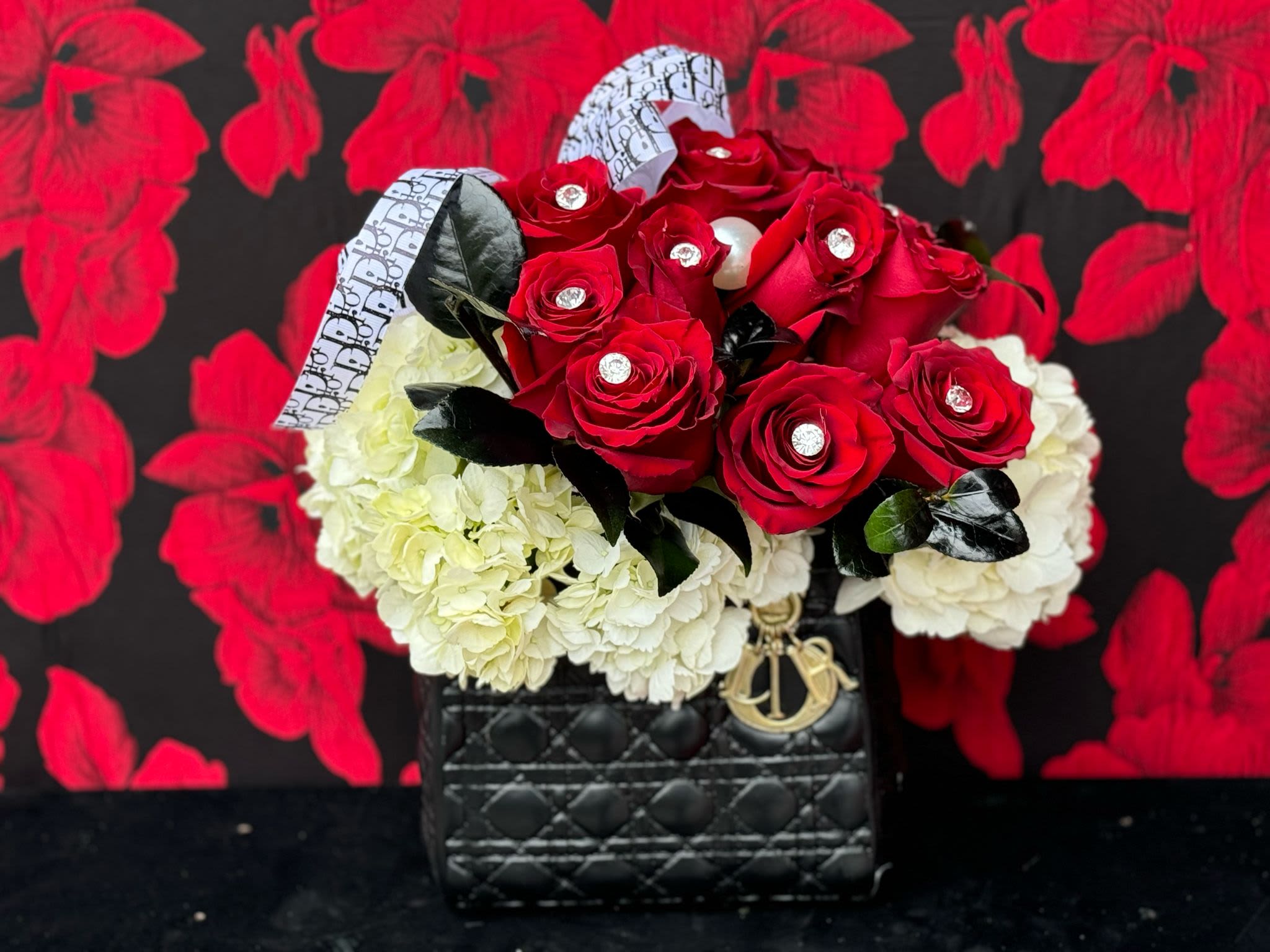 Designer’s Dream - A masterpiece of luxury and style, the "Designer’s Dream" quilted bag is an iconic statement piece fit for a true fashionista. Featuring designer's signature logo, this black  bag exudes elegance and sophistication. Delicate red roses and soft hydrangeas are artfully arranged, adding a soft, romantic touch to the bold, structured design. Subtle diamond details sparkle, bringing an opulent, dazzling finish to this already show-stopping accessory. 