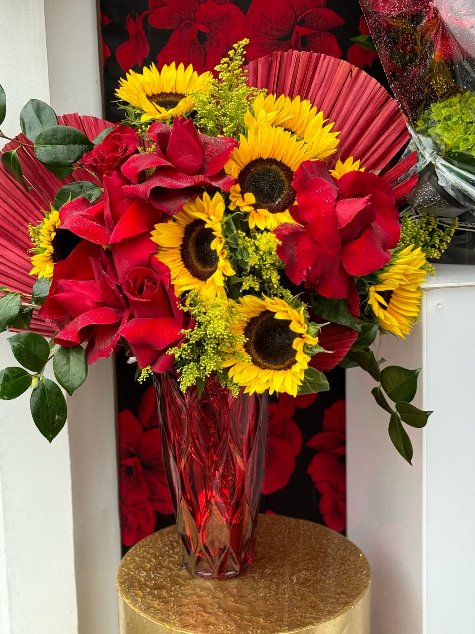 My Sunshine  - A radiant arrangement featuring bright yellow sunflowers paired with rich red roses and complemented by vibrant red palms. The combination of bold florals and lush greenery captures the essence of warmth and positivity, bringing a cheerful and tropical vibe to any space. Making that someone special like a ray of sunshine. 
