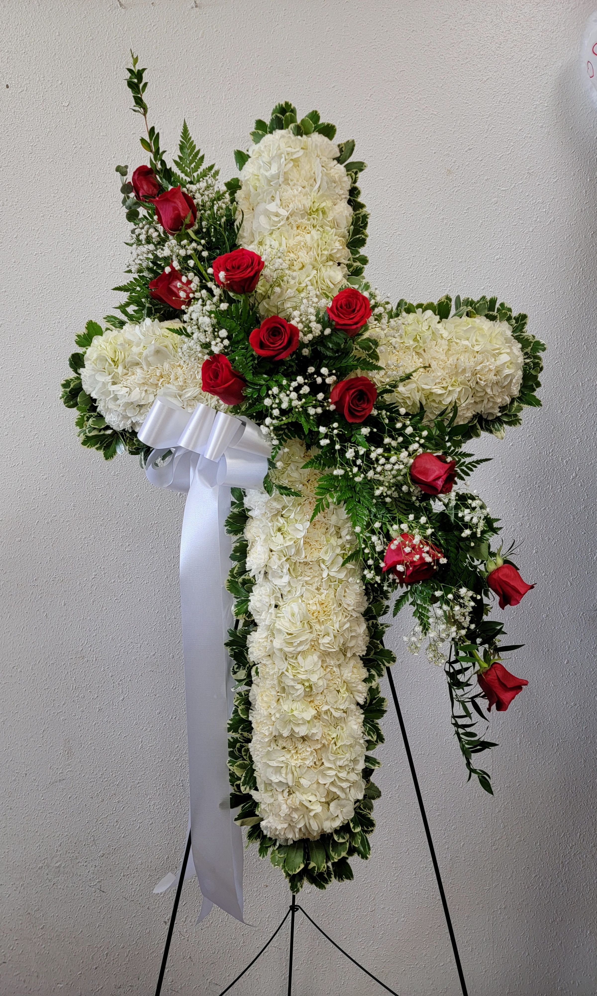 White floral cross with red roses and a white ribbon on a stand
