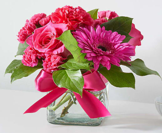 Pink roses, carnations, and a gerbera daisy in a glass vase with a pink ribbon