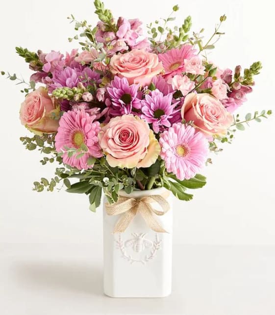 HONEY BEE BUZZ - A soft, garden-style bouquet of blush pink roses and pink daisy-like blooms creates an effortlessly romantic birthday bouquet or anniversary arrangement that will make the person you're thinking of feel cherished. The HONEY BEE BUZZ color palette of pink, white, purple, and green gives a cheerful, springtime impression-perfect for celebrating a milestone, brightening a bedside, or saying "thinking of you" with warmth.  Densely layered blossoms sit in a clean white ceramic vase, accented with airy branching greenery for texture and movement. A natural-toned ribbon tied around the vase adds a delicate finishing touch. The compact, rounded design looks full and abundant on tabletops and mantels; stems are arranged for immediate display, so the recipient can enjoy the soft fragrance and fresh garden feel right away.