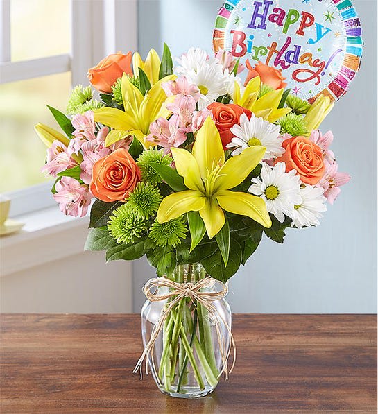 Bouquet of yellow lilies, orange roses, and white daisies in a glass vase with a birthday balloon.