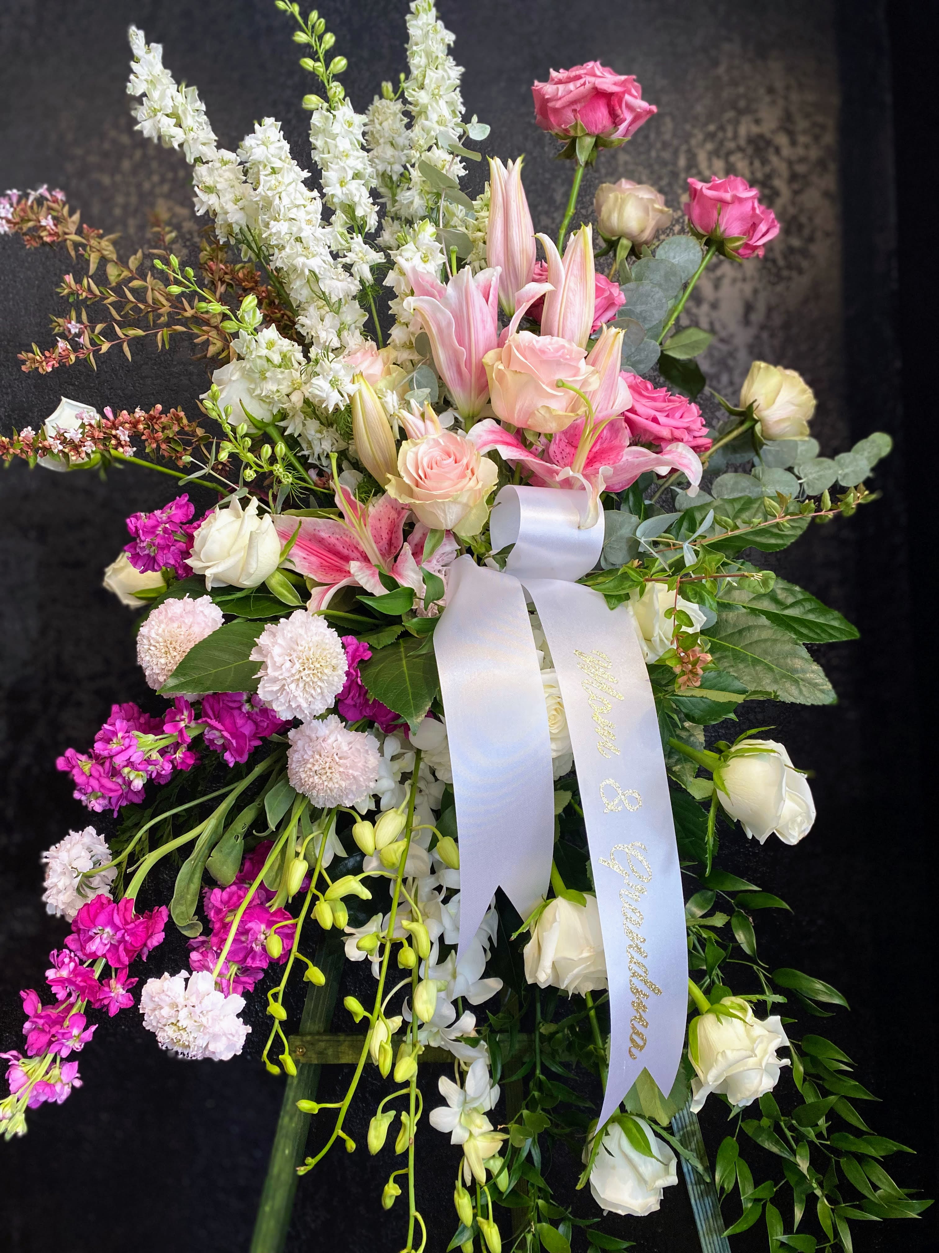 Tall standing spray of pink lilies, roses, and white flowers with a white ribbon on an easel.