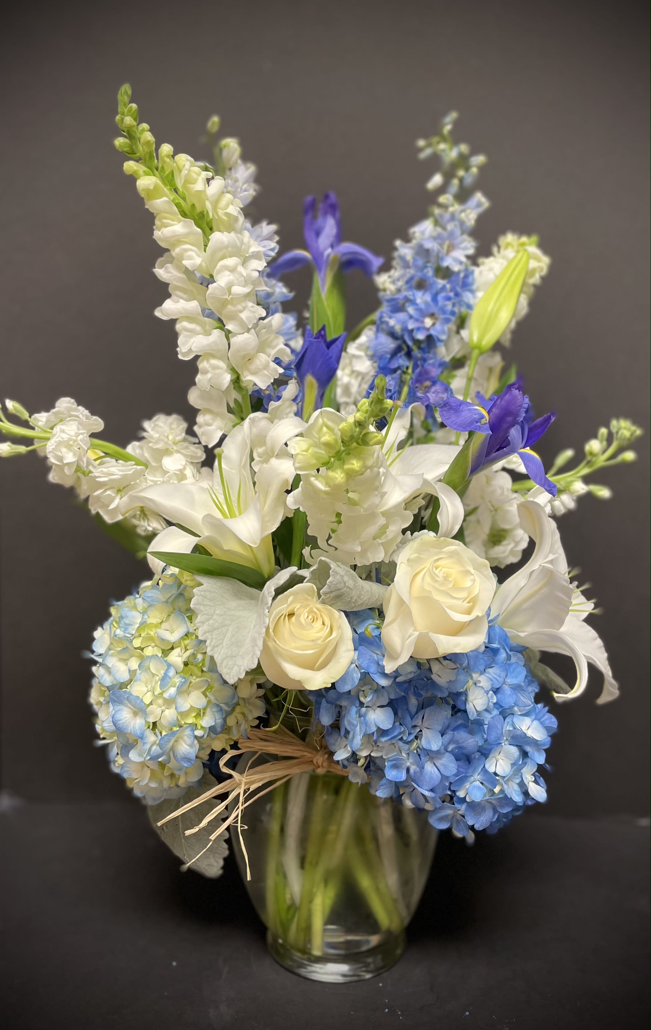 Heaven Sent - White roses, selected light blue hydrangeas, white lilies, blue delphiniums, white stocks, white snapdragons, blue irises, and dusty miller. It comes in a clear ginger vase. All around.