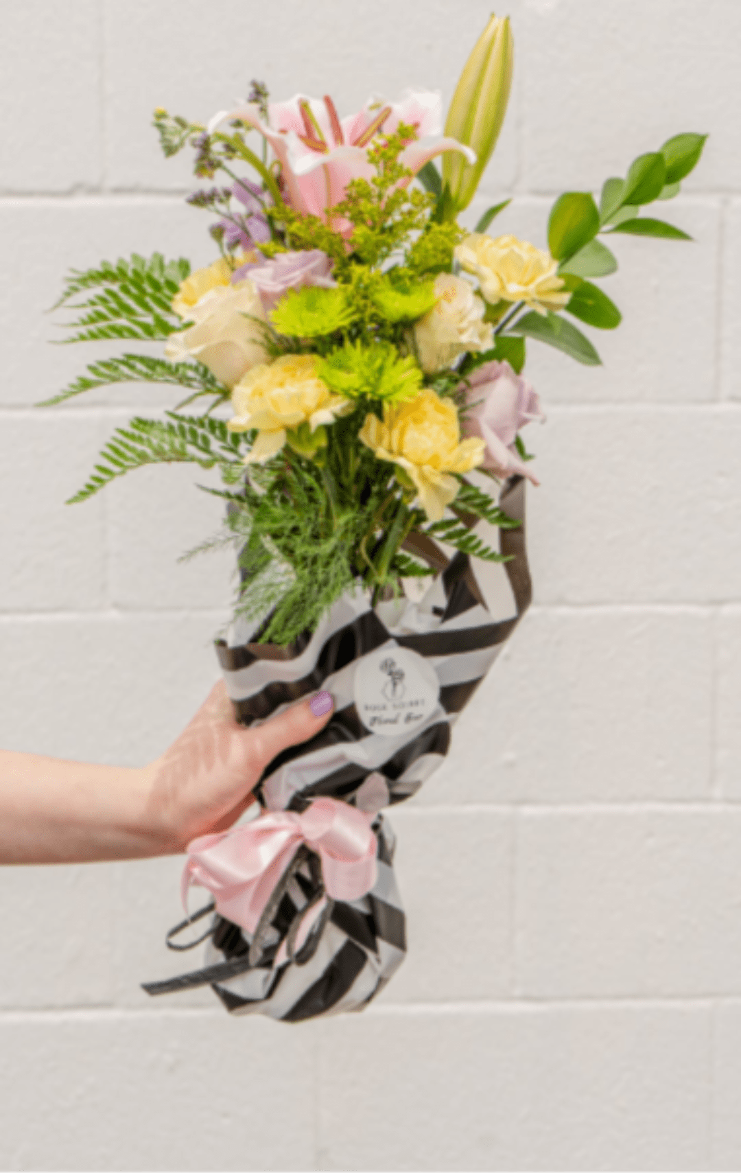 Hand-tied bouquet of pink lilies, yellow carnations, and green mums in striped wrap with a pink bow