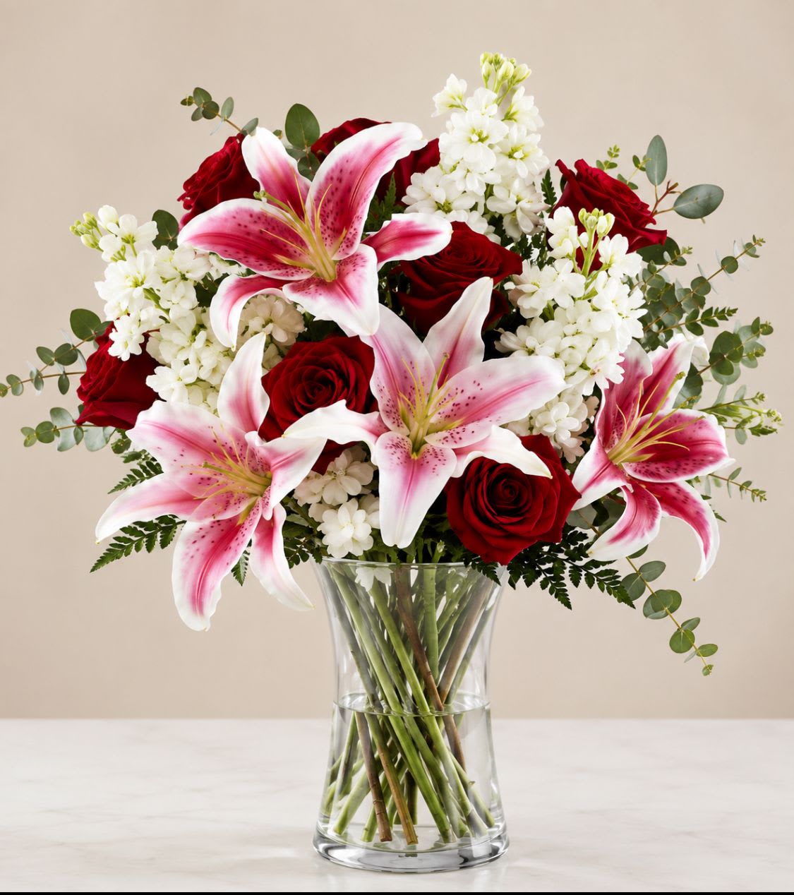 Scarlet symphony - Large pink stargazer lilies unfurl above deep red roses and clusters of crisp white stock in a clear glass vase, making this floral arrangement an immediate statement for anniversaries, love and romance, or a thoughtful sympathy gesture. Scarlet Symphony is designed as a full, rounded bouquet with lilies reaching outward and upward to create height and movement-she'll feel seen and comforted by the lush textures and rich color contrasts.  The mix features stargazer lilies, red roses, and white stock accented with fresh greenery, arranged densely for an abundant, polished look. Colors range from saturated crimson through soft pink to clean white, giving the arrangement both dramatic depth and tender softness. Presented in a simple clear glass vase, the display measures as a centerpiece-sized arrangement suitable for tabletops, bedside stands, or the front of a mantle.  Perfect for expressing "I'm sorry," offering condolences, marking a graduation, or celebrating a wedding or anniversary, this bouquet sends a message of heartfelt care. For best longevity, place in fresh water, trim stems slightly when refreshing the vase, and keep away from direct heat; ordering options include delivery for special dates like Valentine's Day, Mother's Day, or Women's Day to ensure timely arrival.