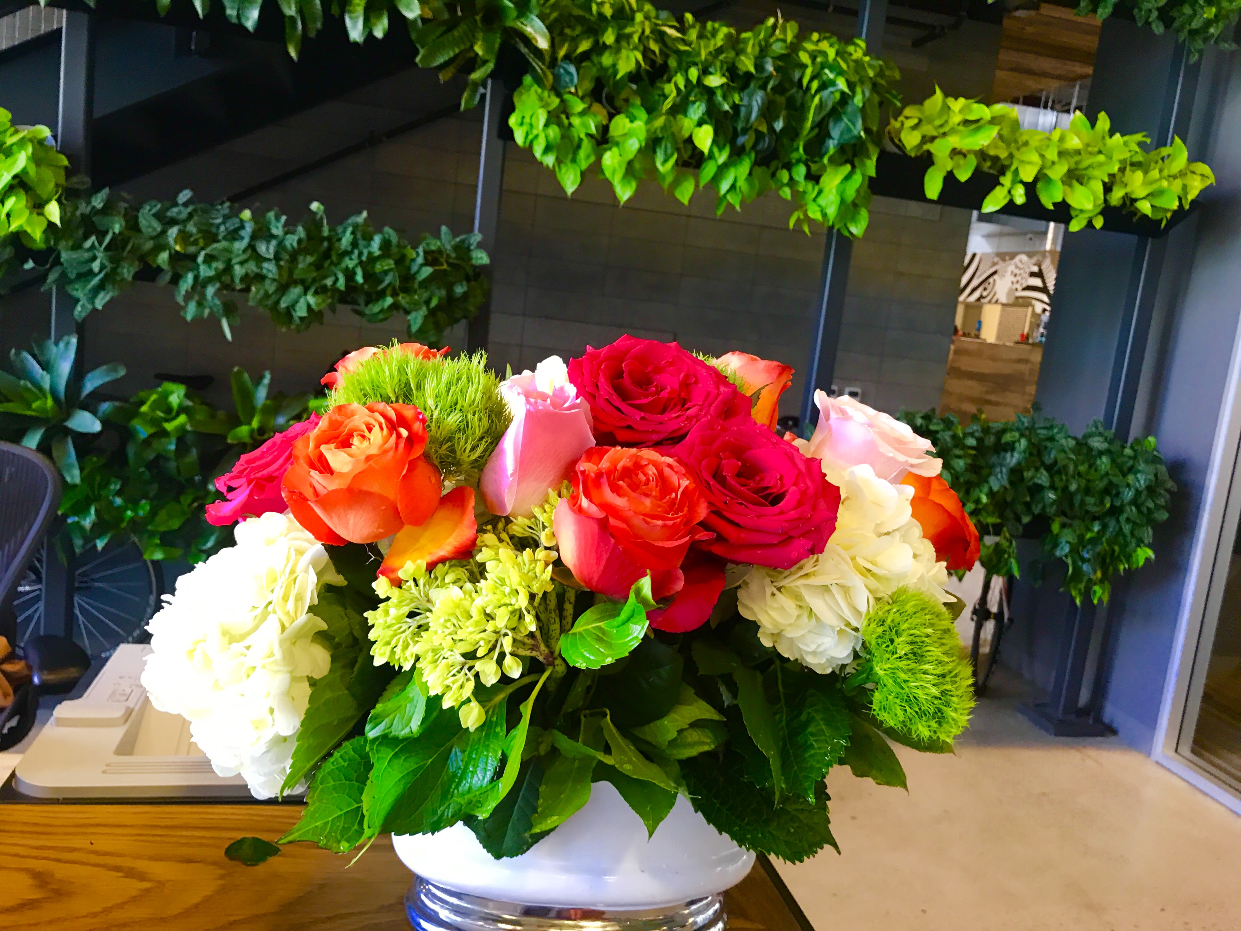 Low arrangement of red, orange, and pink roses with white hydrangeas in a white bowl vase