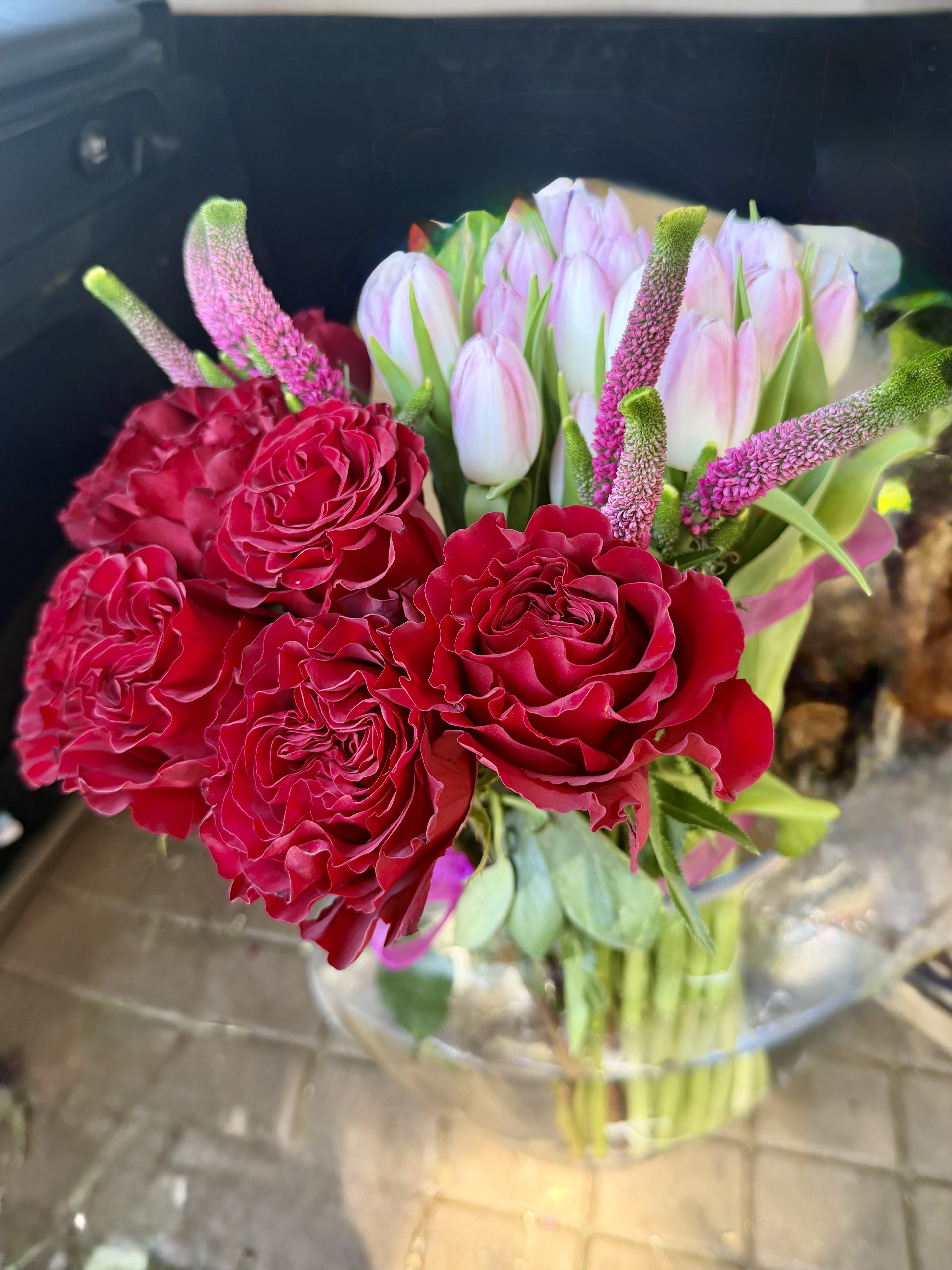 Hydrangeas , tulips ,red rose arrangement - Velvety red roses take center stage in this red rose arrangement, surrounded by pale pink tulips and airy white-and-green accents to create a romantic anniversary or Valentine's Day bouquet. The mix of saturated red and soft blush tones reads both celebratory and intimate, the kind of floral gift that makes the person you're thinking of feel truly cherished.  Arranged with a slightly modern, hand-tied look and presented in a clear glass vase with decorative wrap, the design pairs large, lush roses with upright pink-and-green spike flowers for height and texture. White blooms and fresh greenery weave through the composition to brighten the palette and add visual contrast, making it equally lovely for spring gifting.  Perfect for anniversaries, Valentine's Day, or a springtime surprise, this bouquet arrives ready to display in the included vase; just trim stems and refresh water as needed to keep blooms at their best. The combination of classic roses and seasonal tulips sends a timeless message of affection and celebration.