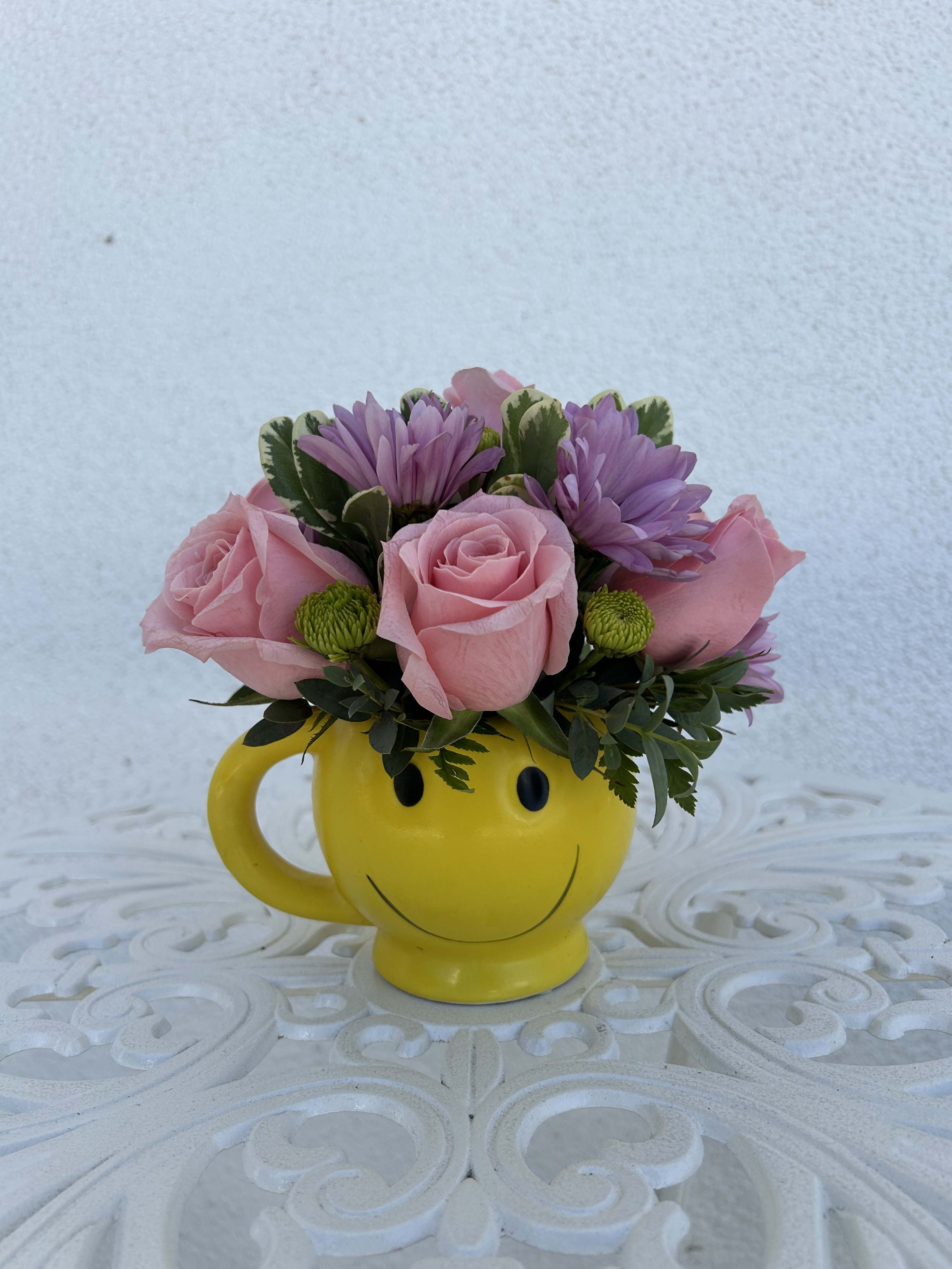Pink roses and purple daisies arranged in a yellow smiley-face mug